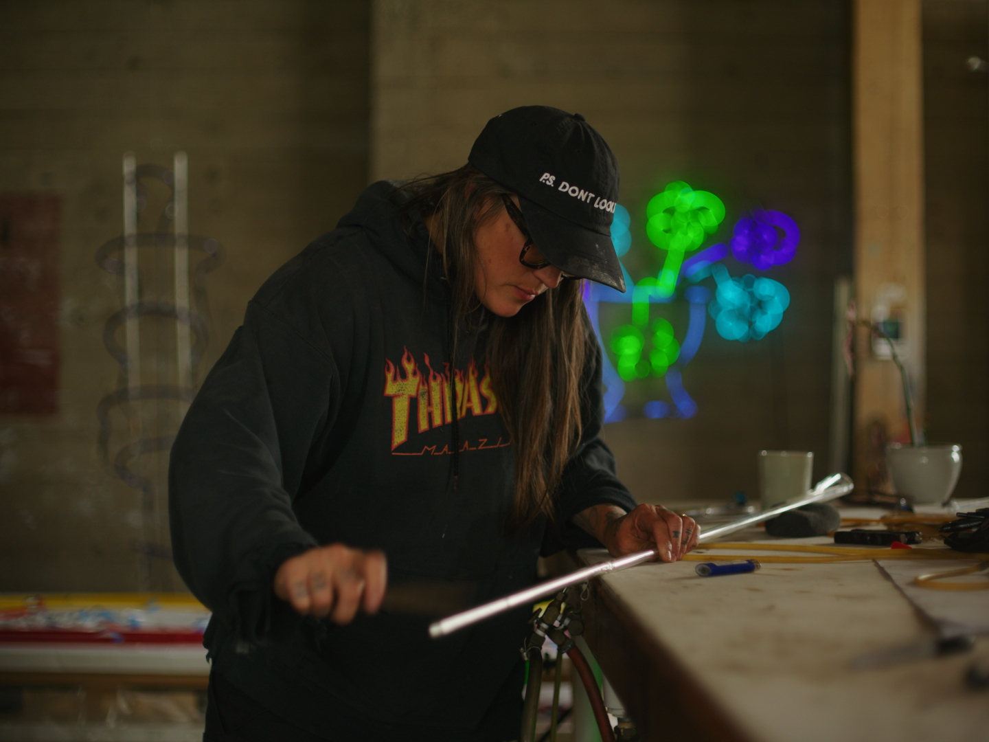 A woman wearing a black hat and hoodie working on a glass piece at a workbench in a dimly lit studio with colorful neon art on the wall behind her.