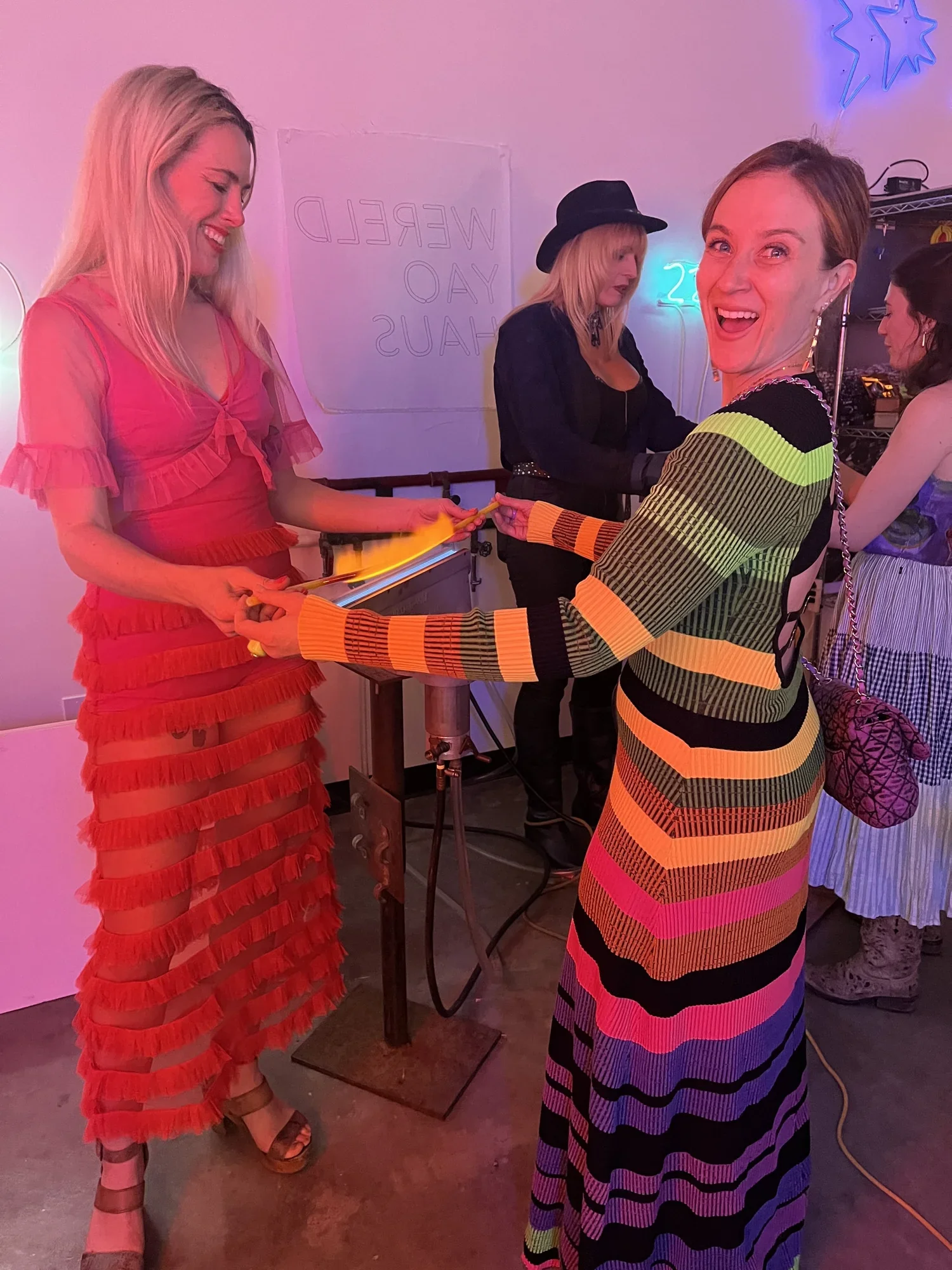 Group of women at a vibrant party, exchanging a gift, with neon signs and colorful lighting.