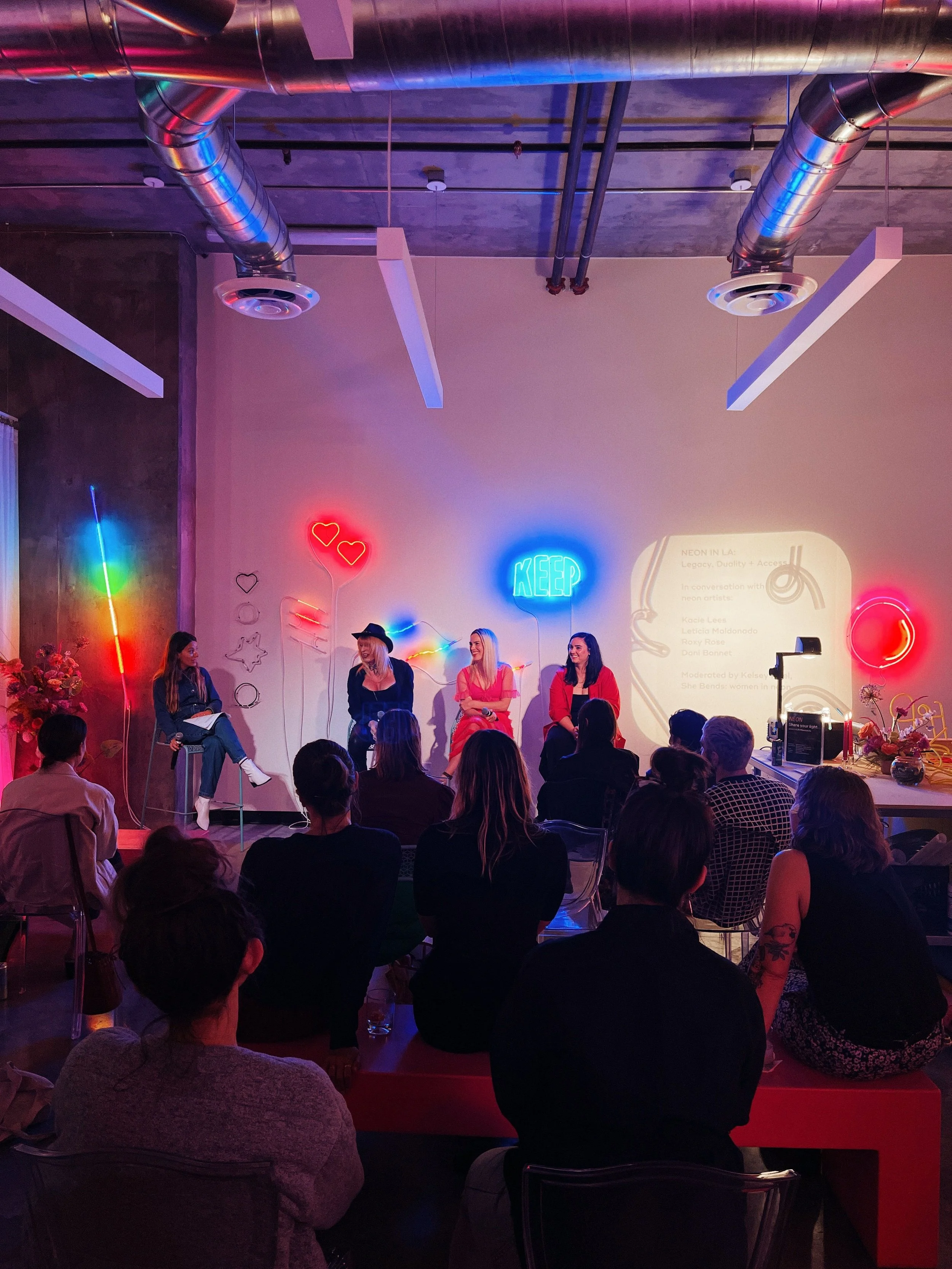 Indoor event with four women panelists sitting on stage with neon signs reading 'KEEP' and hearts behind them, and an audience seated facing the stage.