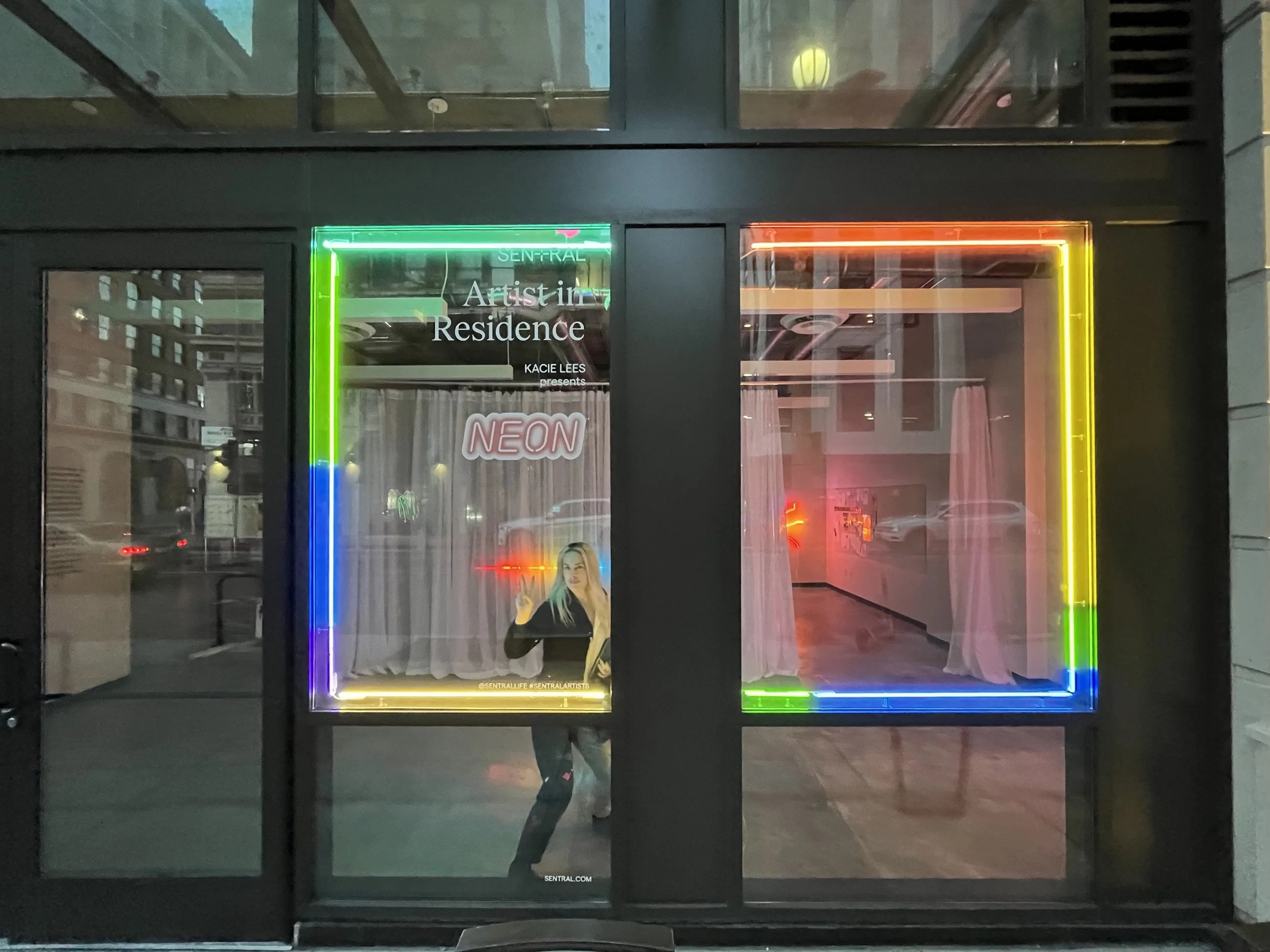 Neon sign on window advertising 'Artist in Residence' with rainbow-colored border, a person giving a peace sign inside, and curtains in the background.