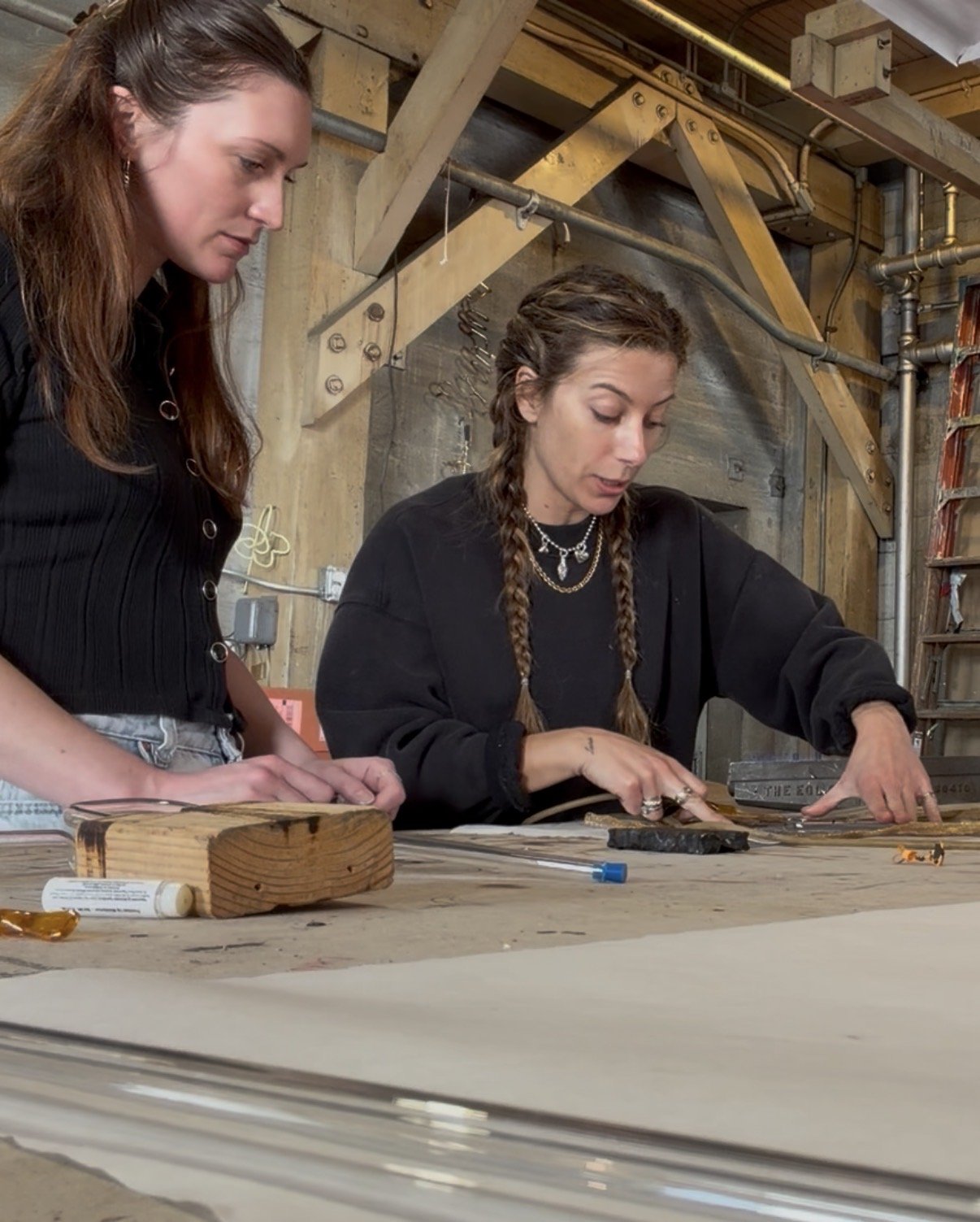 Two women are working on a project at a wooden table in a workshop with exposed beams and pipes. One woman with braids is pointing at something on the table, while the other woman observes.