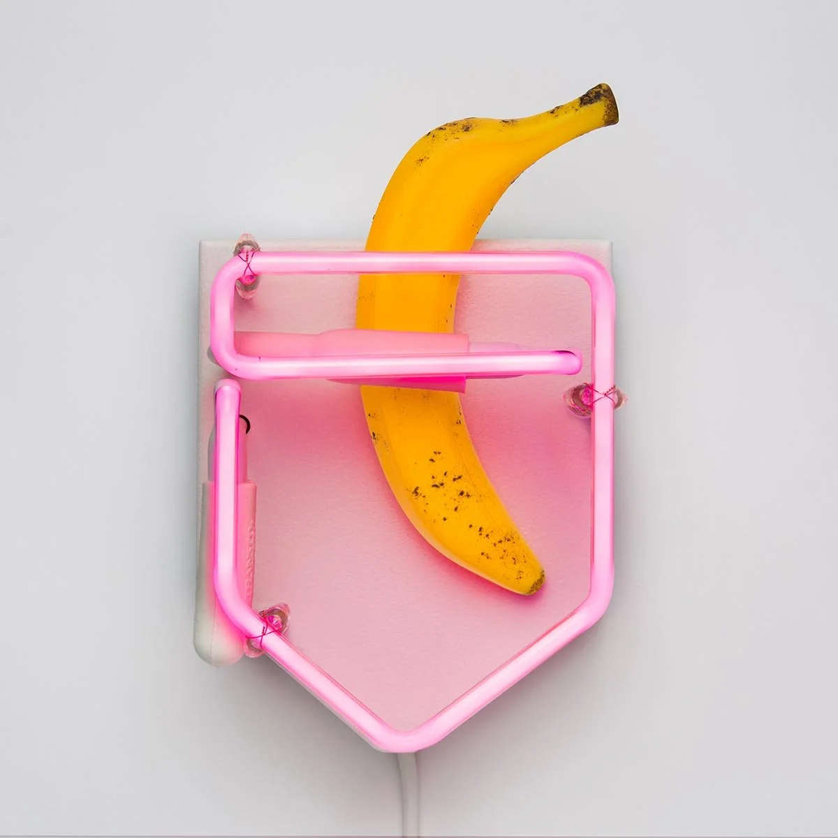 A yellow banana with brown spots inside a pink neon sign shaped like a shield on a white background.