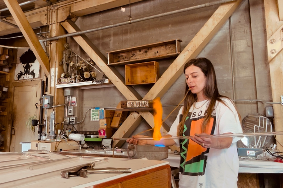 A woman working with glass blowing equipment in a workshop, with a lit piece of molten glass on a metal tube.