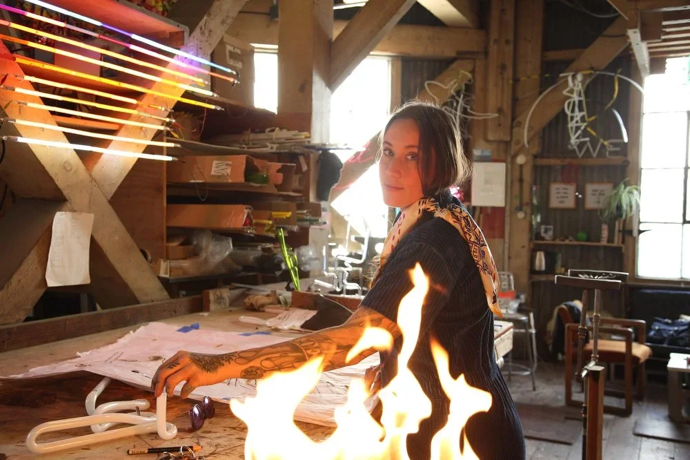 Young woman with a tattooed arm working at a cluttered wooden workbench in a rustic studio, with fire in front of her and neon lights on the wall.