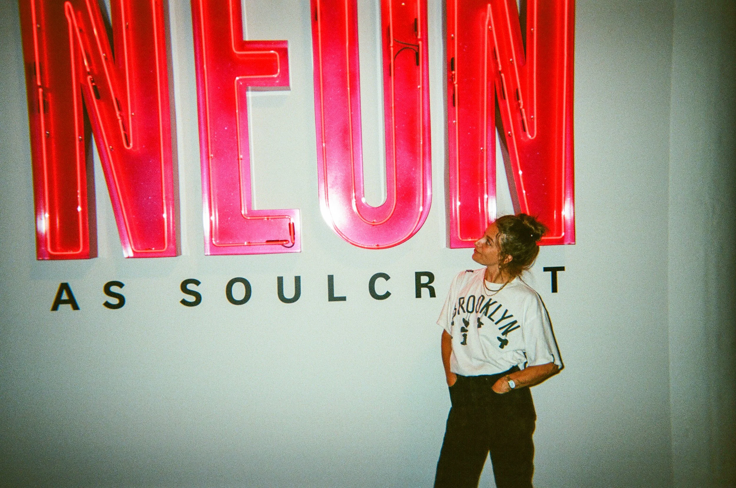 A woman standing in front of a wall with a large pink neon sign that reads "NEON." Below the sign, there are black letters spelling "AS SOULC." She is wearing a white Brooklyn t-shirt, black pants, and has her hair tied up in a bun.