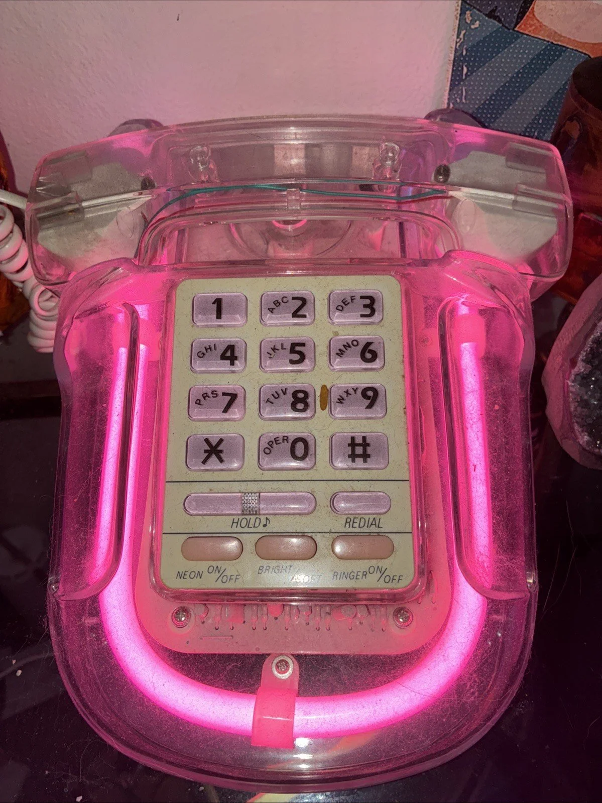 Pink neon-lit vintage telephone with a transparent outer casing and a numeric keypad, resting on a dark surface.