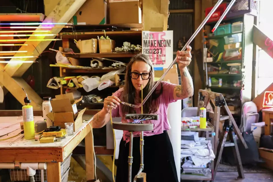 A woman with glasses and tattoos wearing a pink shirt, she is blowing glass in a workshop filled with tools, supplies, and colorful objects.