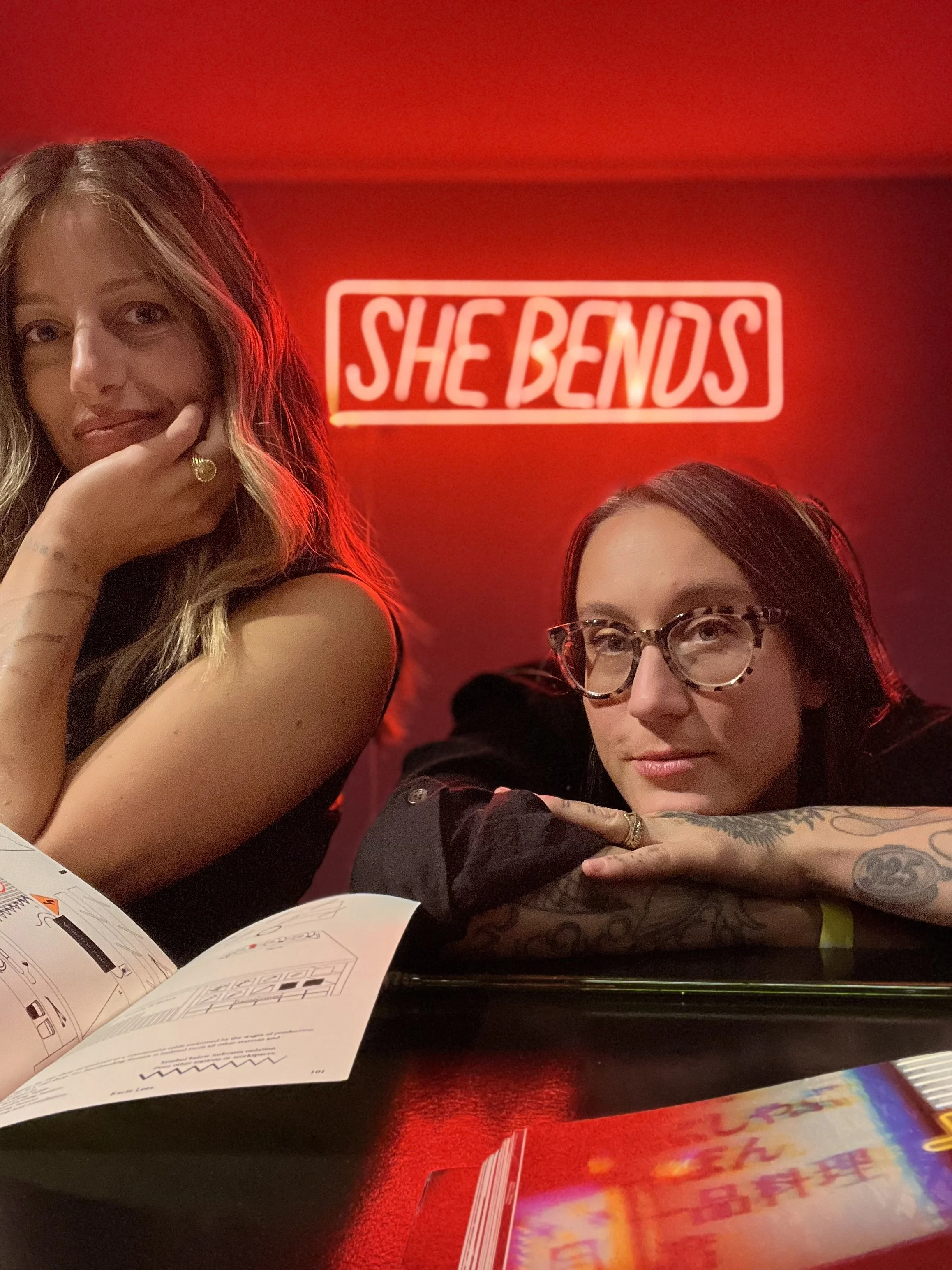 Two women sitting at a table with a neon sign that says 'She Bends' behind them, one resting her chin on her hand and the other leaning on her arm, with various items on the table in front of them.