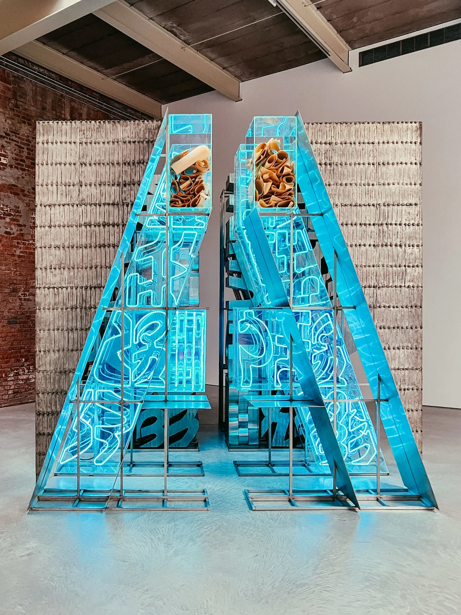 Art installation featuring two large blue neon letter A's with neon piping inside, standing on a concrete floor in an industrial-style gallery with brick and textured wall background.