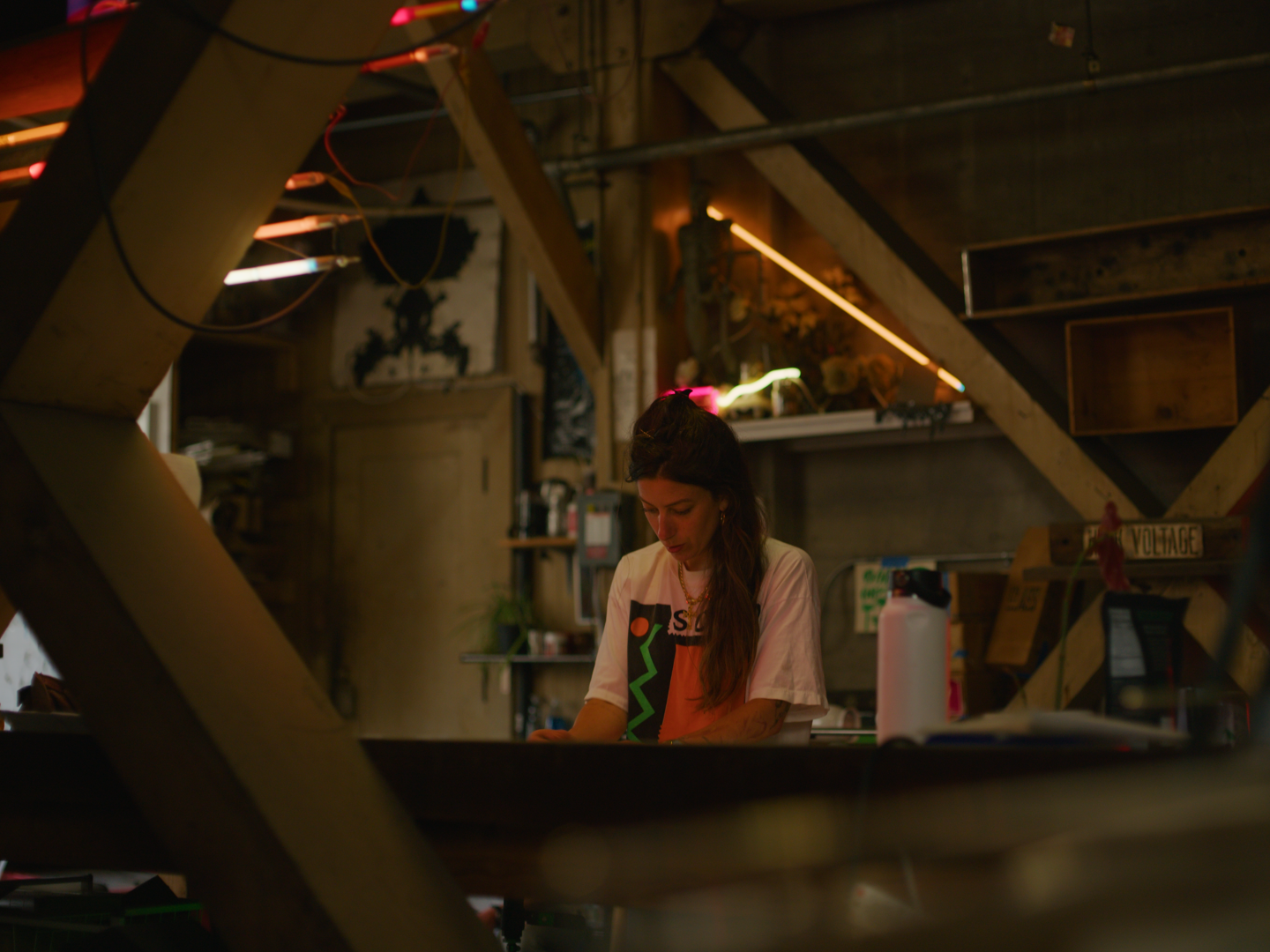 A woman with long dark hair working at a glass bending table in an industrial-style neon studio with exposed wood beams and shelves.