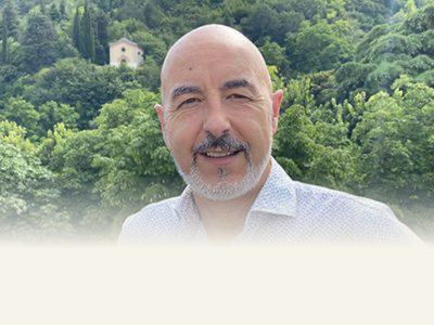 A smiling man with a bald head and a beard standing outdoors with a lush green forest and a small house in the background.