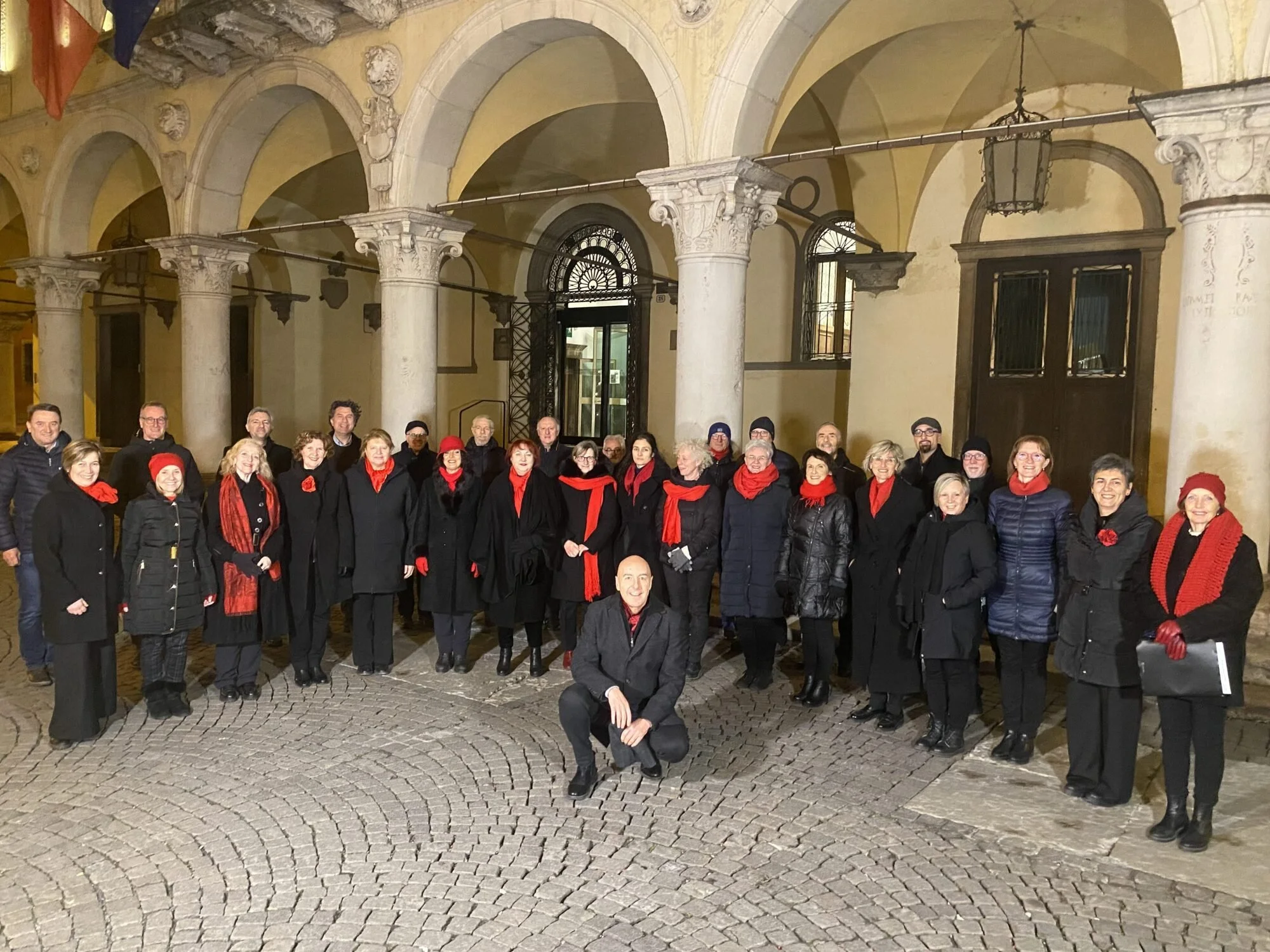 A group of people dressed in black with red accessories, gathered outside in front of arched columns and historic architecture at night.