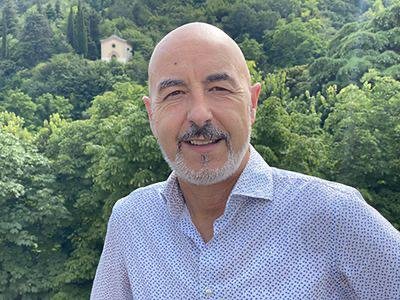 A smiling middle-aged man with a beard and bald head, wearing a light blue patterned shirt, standing outdoors with green trees and a small white building in the background.