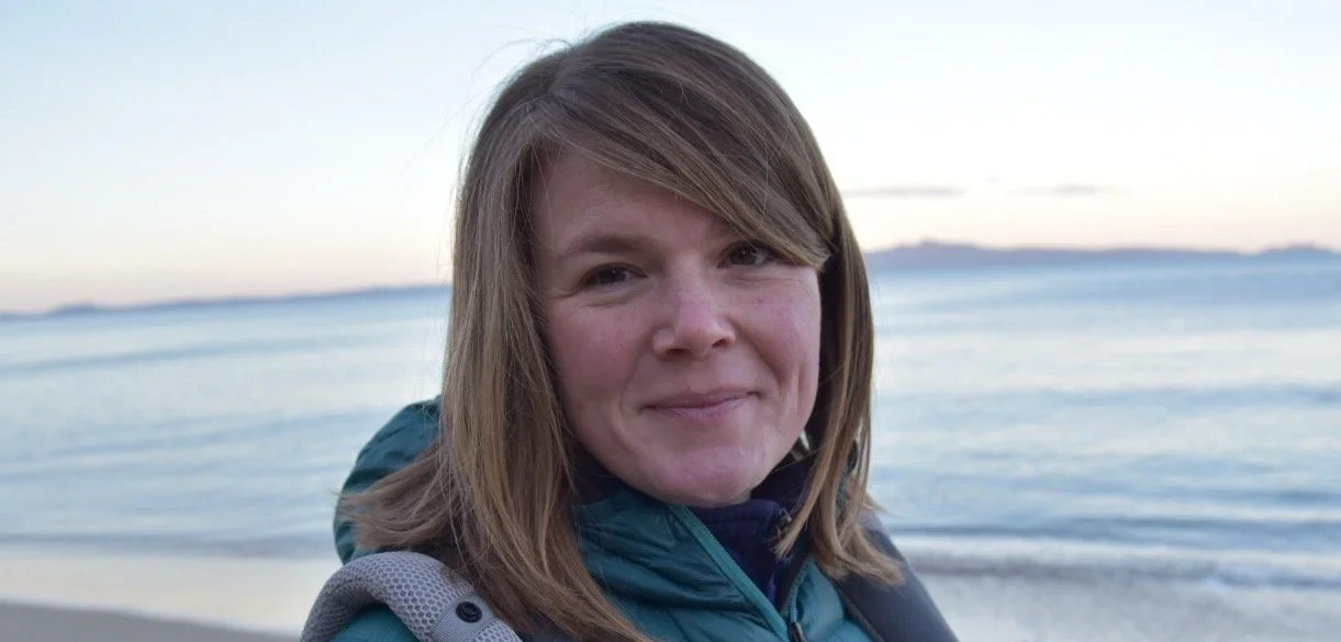 A woman with shoulder-length brown hair smiling at the camera on a beach during sunset, with water and distant land in the background.