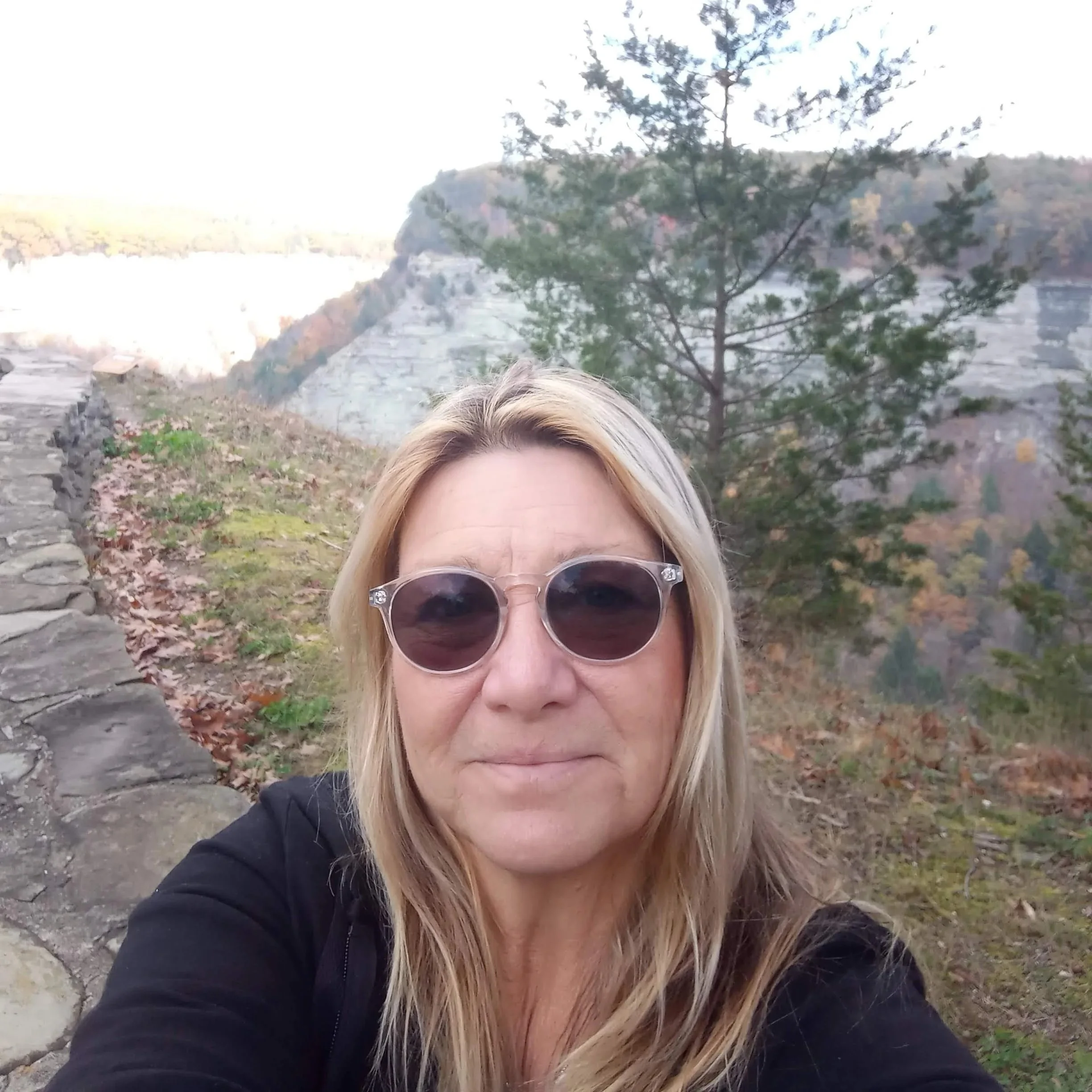 Close-up of a woman with blonde hair wearing sunglasses and a black jacket, outdoors near a scenic landscape with trees, rocky path, and a canyon in the background.