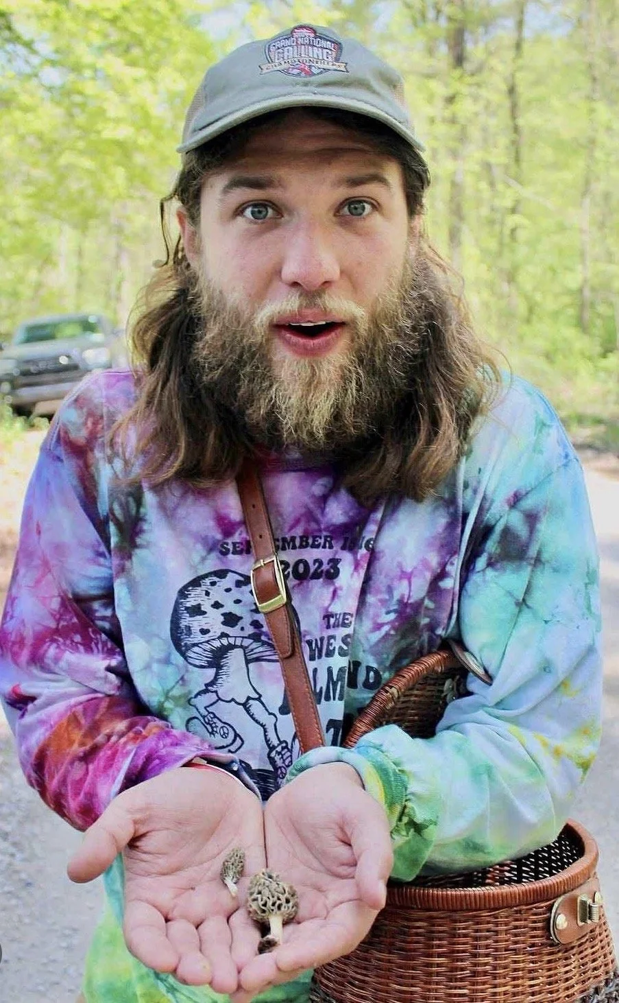 A man with long hair and a beard wearing a colorful tie-dye shirt and a gray cap, holding small mushrooms in his hands. He is outdoors in a wooded area with a car in the background.