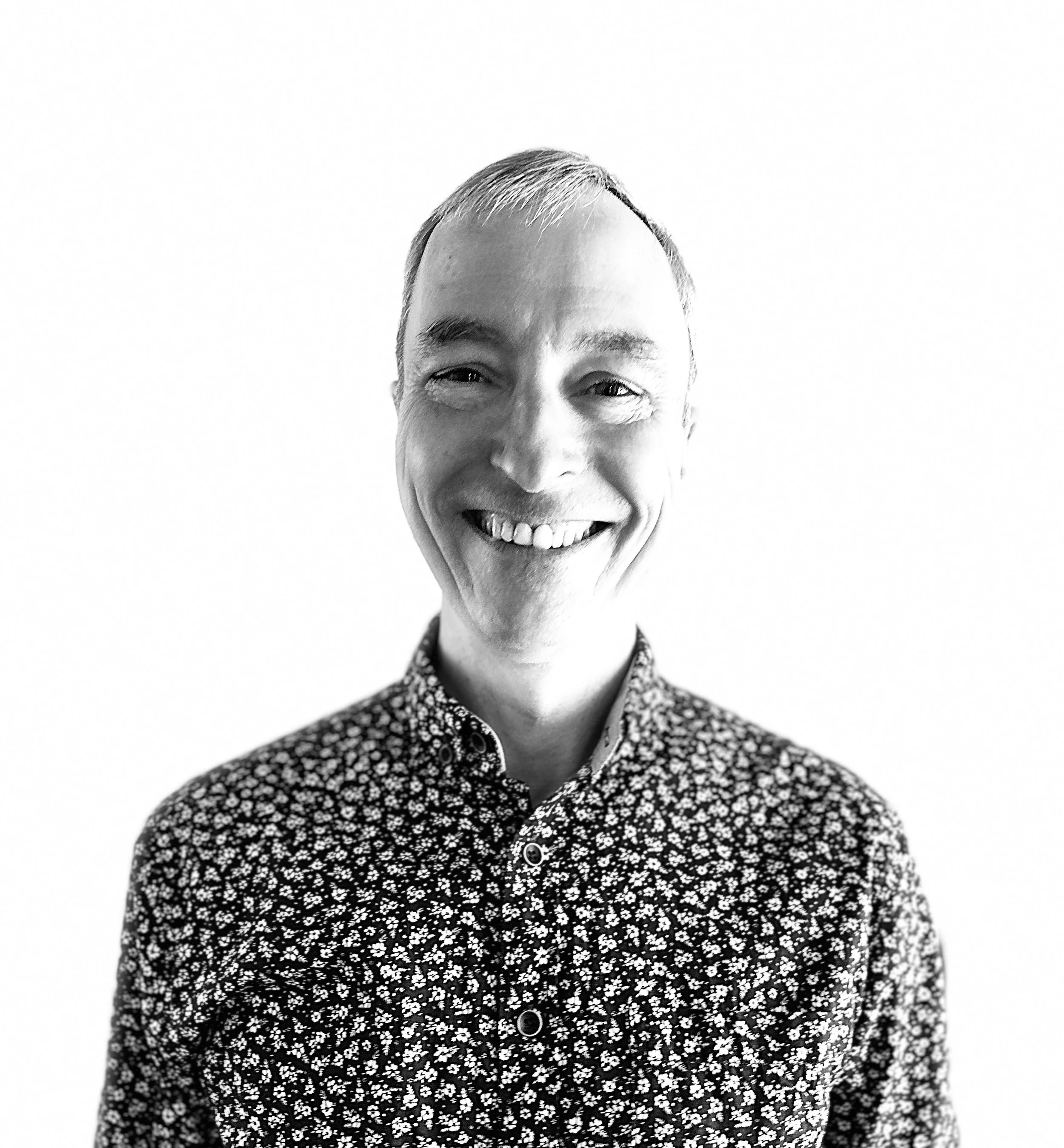 Portrait en noir et blanc d'un homme souriant portant une chemise à motifs floraux.