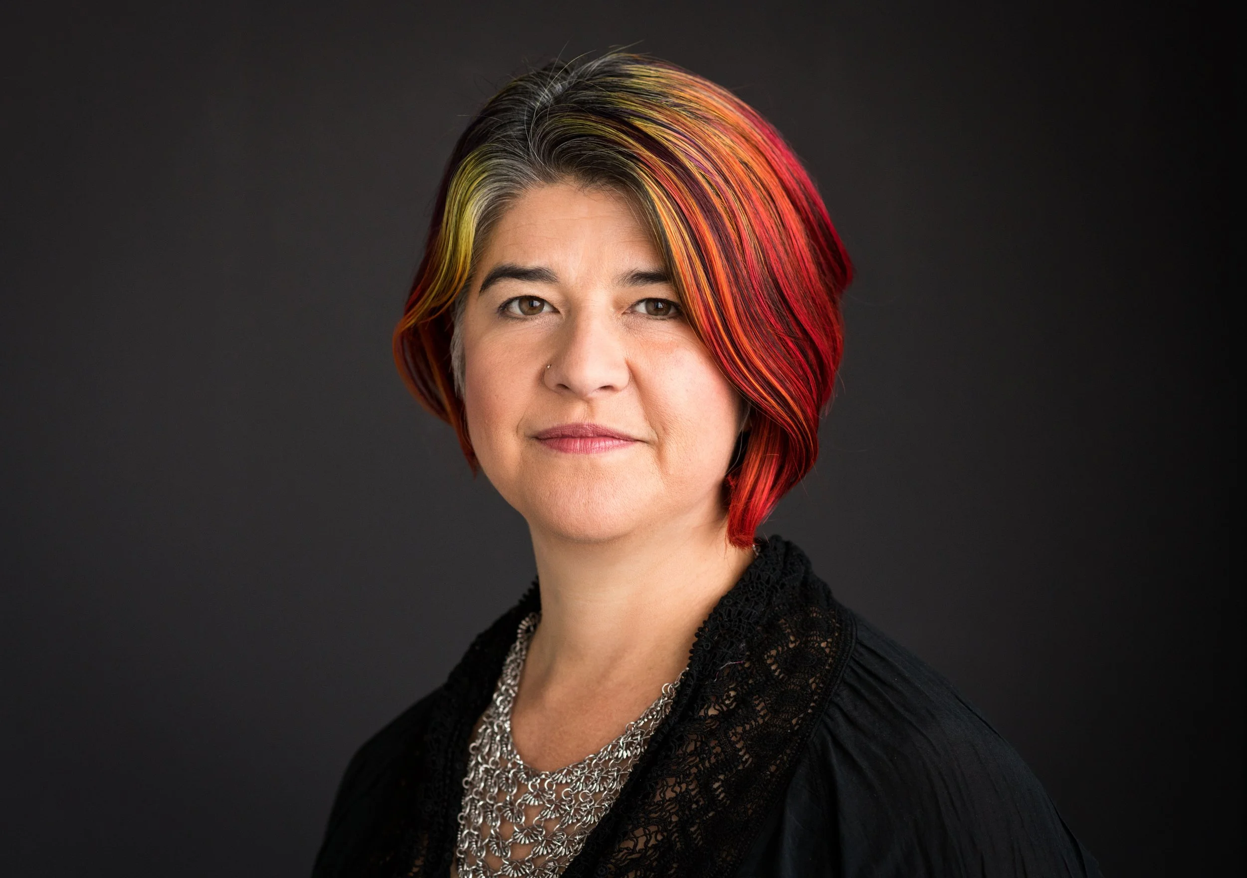 Portrait of a woman with short hair featuring multicolored highlights of yellow, orange, and red, wearing a black top and a silver chain necklace, against a dark background.