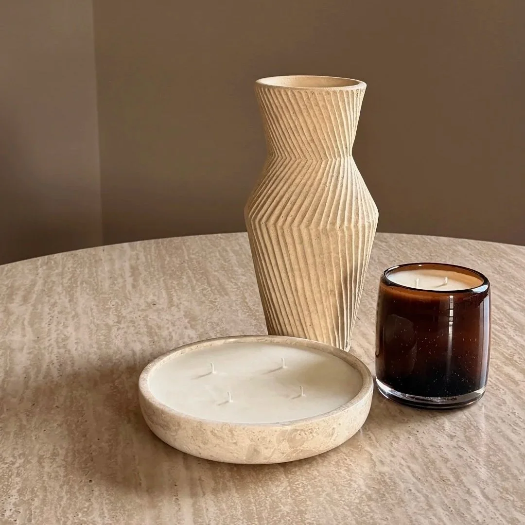 Decorative table display with a beige textured vase, a round stone candle holder with no candles, and a small dark brown glass candle holder with an unlit candle on a light-colored surface.