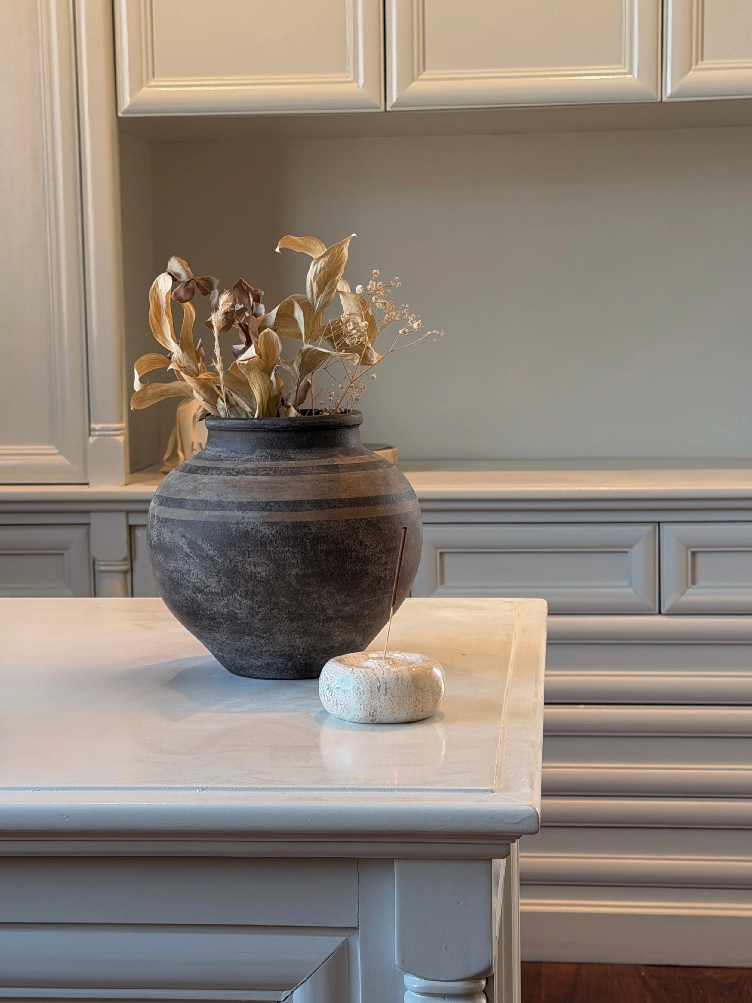 A ceramic vase with dried leaves and flowers on a white tabletop, with kitchen cabinetry in the background.