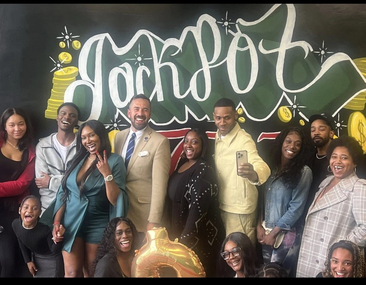 A group of diverse people posing and smiling in front of a chalkboard with the words 'jackpot' and dollar signs, celebrating together.