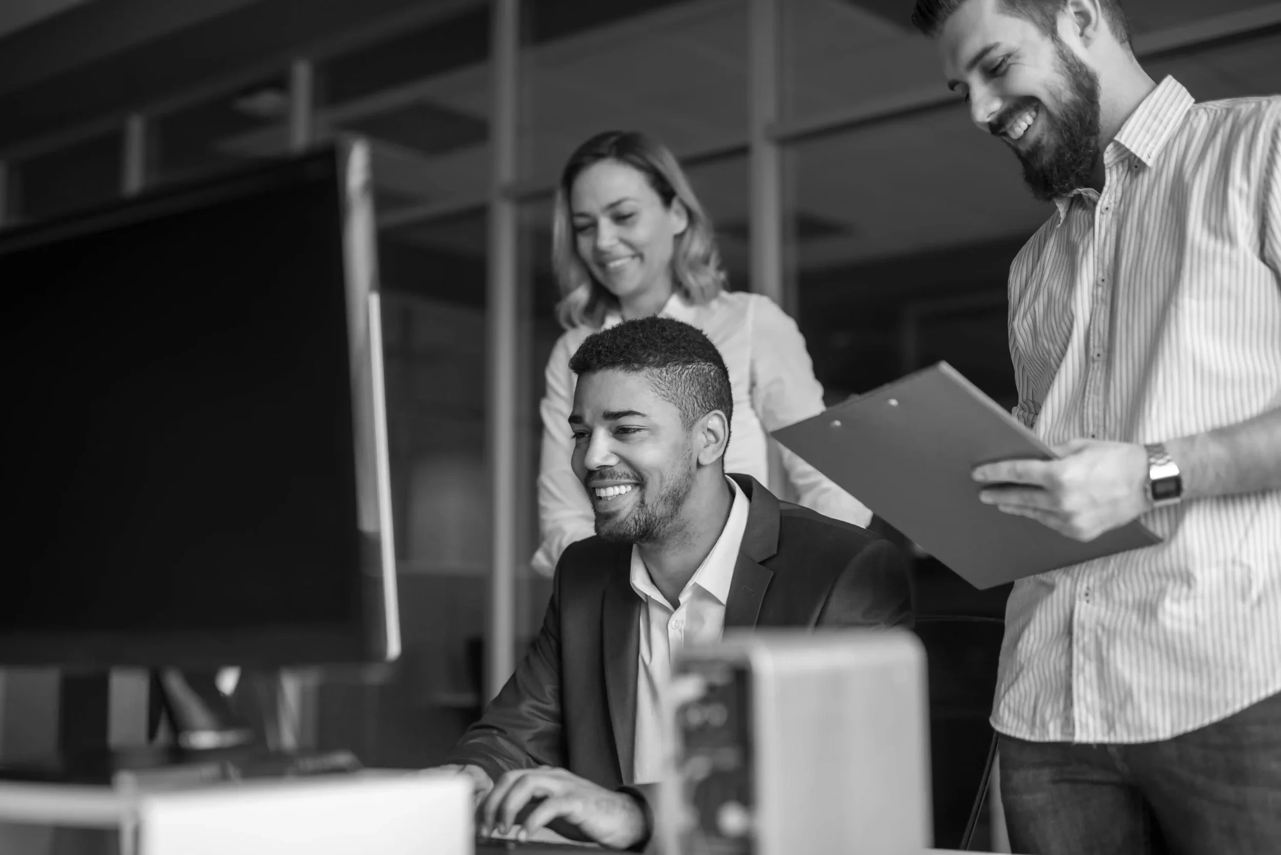 Three colleagues in an office looking at a computer screen, with one seated man and two standing women, all smiling, discussing work.