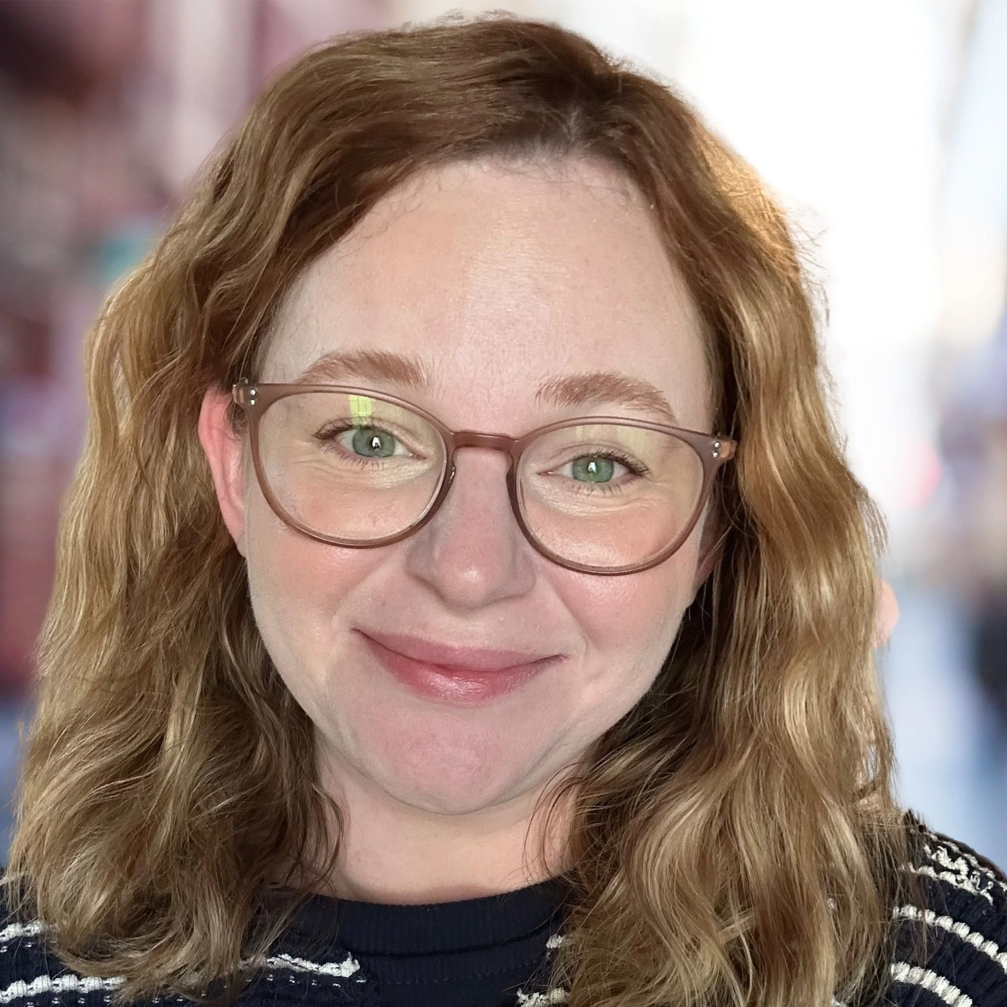 Close-up of a smiling woman with wavy red hair, wearing glasses, and a dark striped shirt, with a blurred urban background. Therapist. Licensed clinical social worker.
