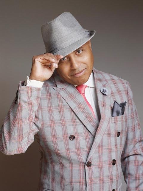 A man wearing a gray fedora hat, a plaid suit jacket, a white shirt, and a pink tie, tipping his hat and smiling.