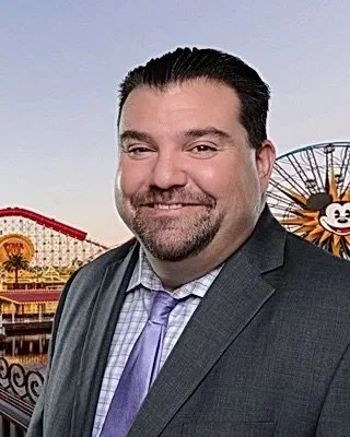 A man in a suit with a tie, smiling, with an amusement park in the background featuring a Ferris wheel and a roller coaster.