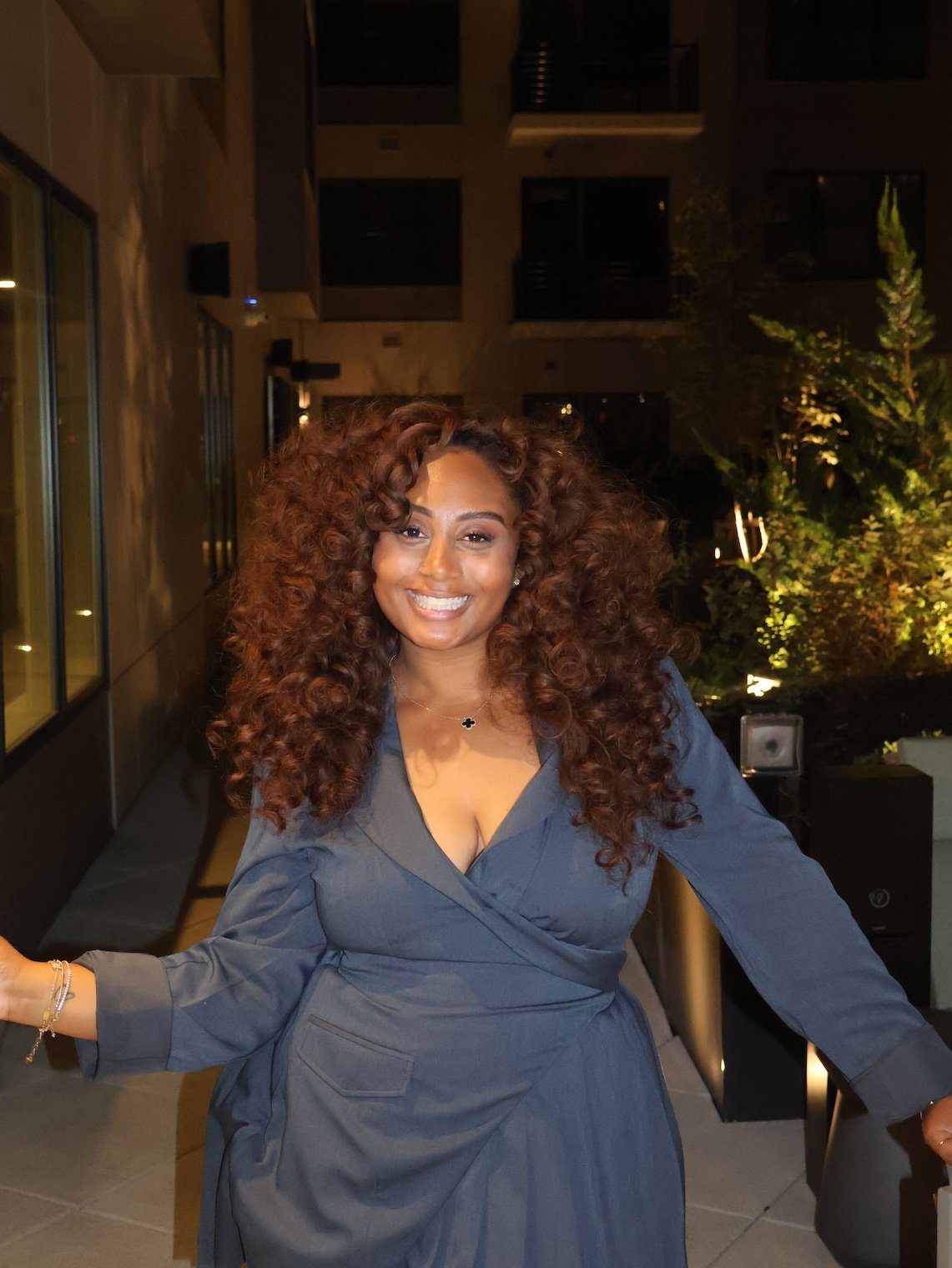 A woman with long, curly brown hair smiling at a nighttime outdoor setting.