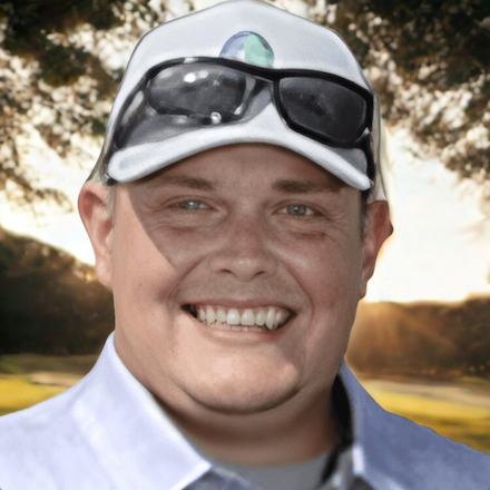 Smiling man wearing sunglasses and a white hat with a lens on top, outdoors with trees and a sunset background.