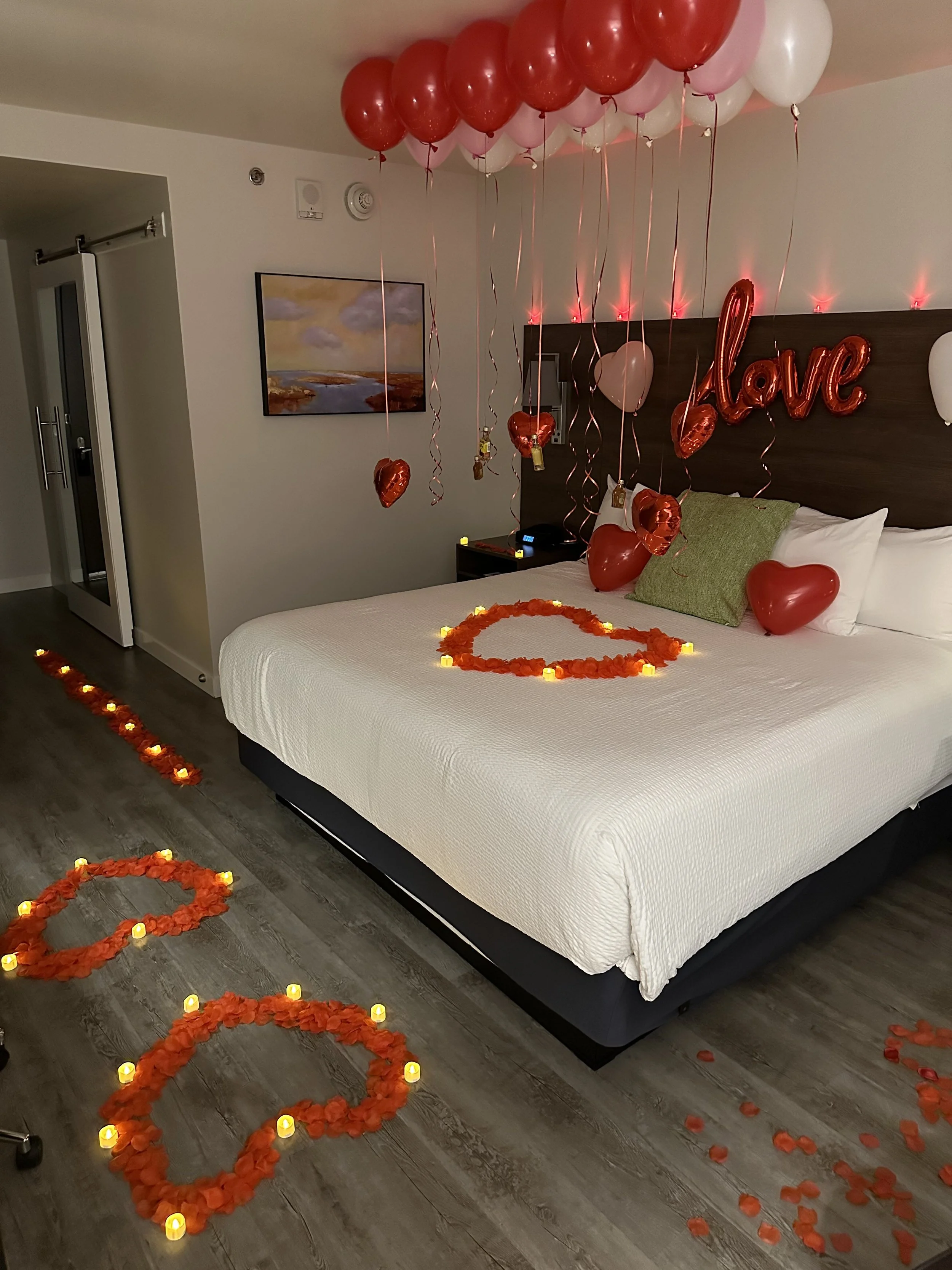 A decorated hotel room with red and white balloons, heart-shaped balloons, and LED candles arranged in heart shapes and on the floor, creating a romantic atmosphere for Valentine's Day.