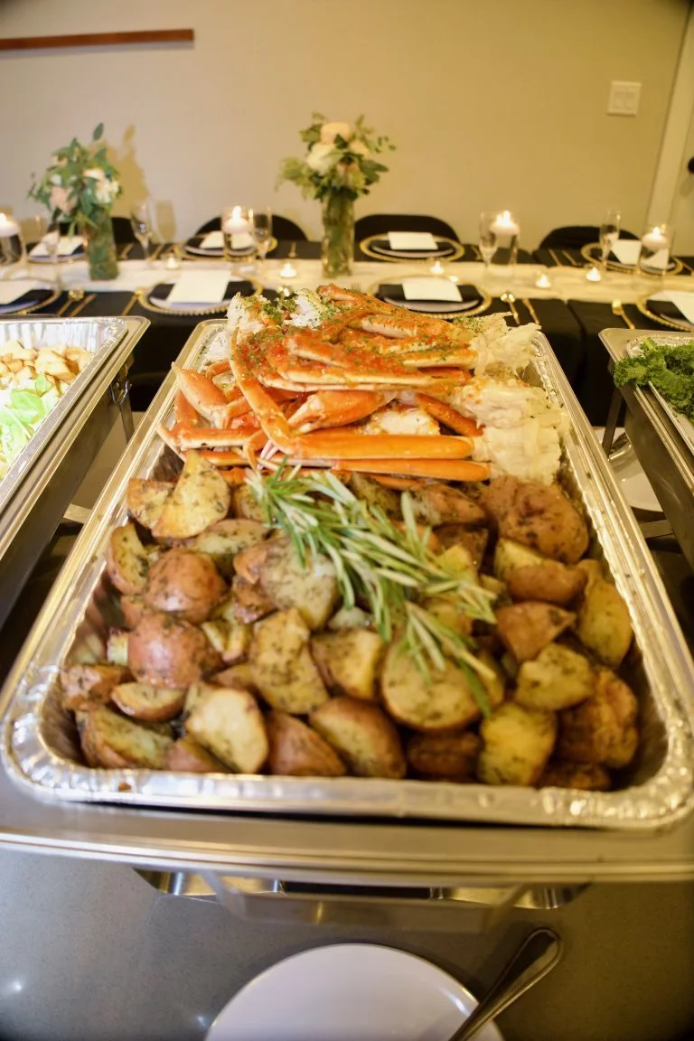 Buffet tray of roasted potatoes garnished with rosemary, and cooked crab legs topped with herbs, in a dining setting with a table decorated with white flowers and candles.