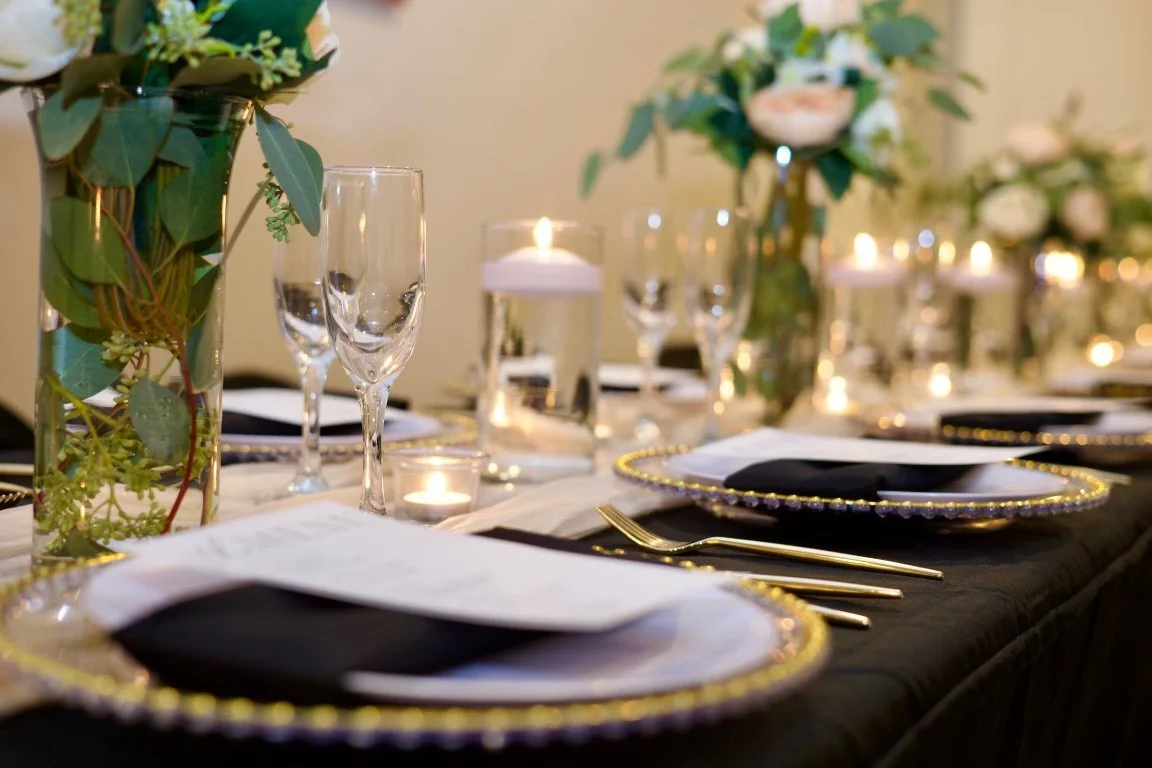Elegant dining table with black and gold-rimmed plates, gold flatware, water glasses, candles, and floral centerpieces with greenery and white flowers.