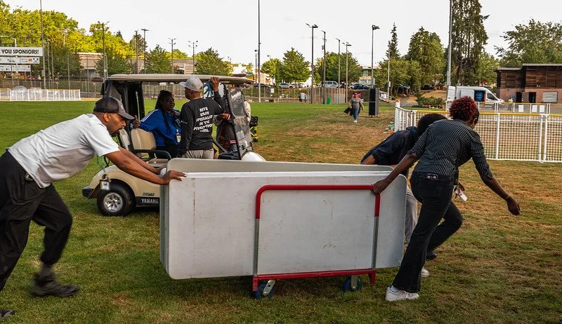 Group of people moving a large white object, possibly a wall or partition, across a grassy field near a golf cart, with others in the background near a fenced sports field.