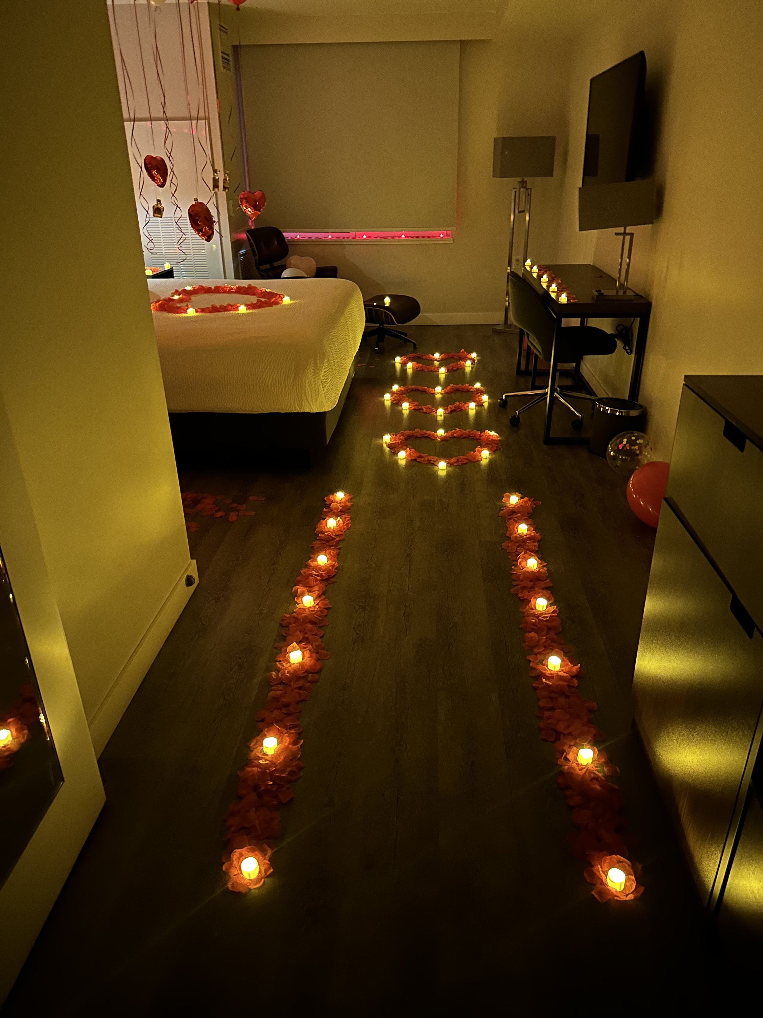 A dimly lit room decorated with Valentine's Day themed items. Two lines of candles with flowers on top form a pathway leading to a bed with a heart-shaped arrangement of candles and flowers. Heart-shaped hanging balloons and Valentine's Day decoratio