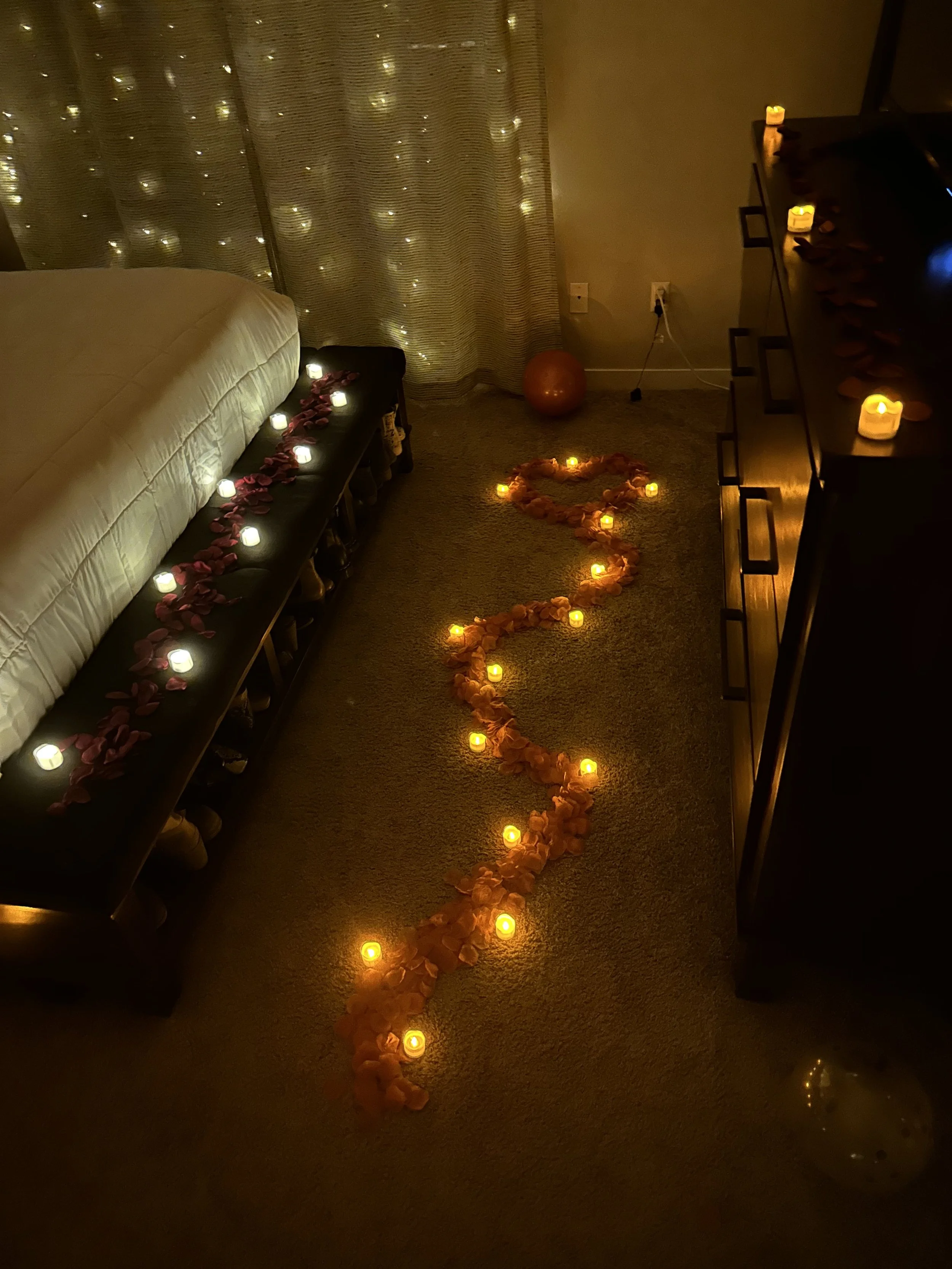 Romantic bedroom decorated with candles and rose petals, with a string of lights on the floor forming a heart shape.