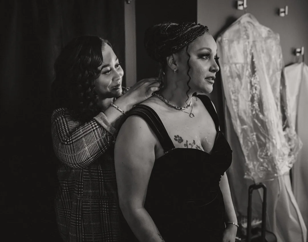 A woman is being helped with her dress by another woman in a bridal shop, with wedding dresses hanging in the background.