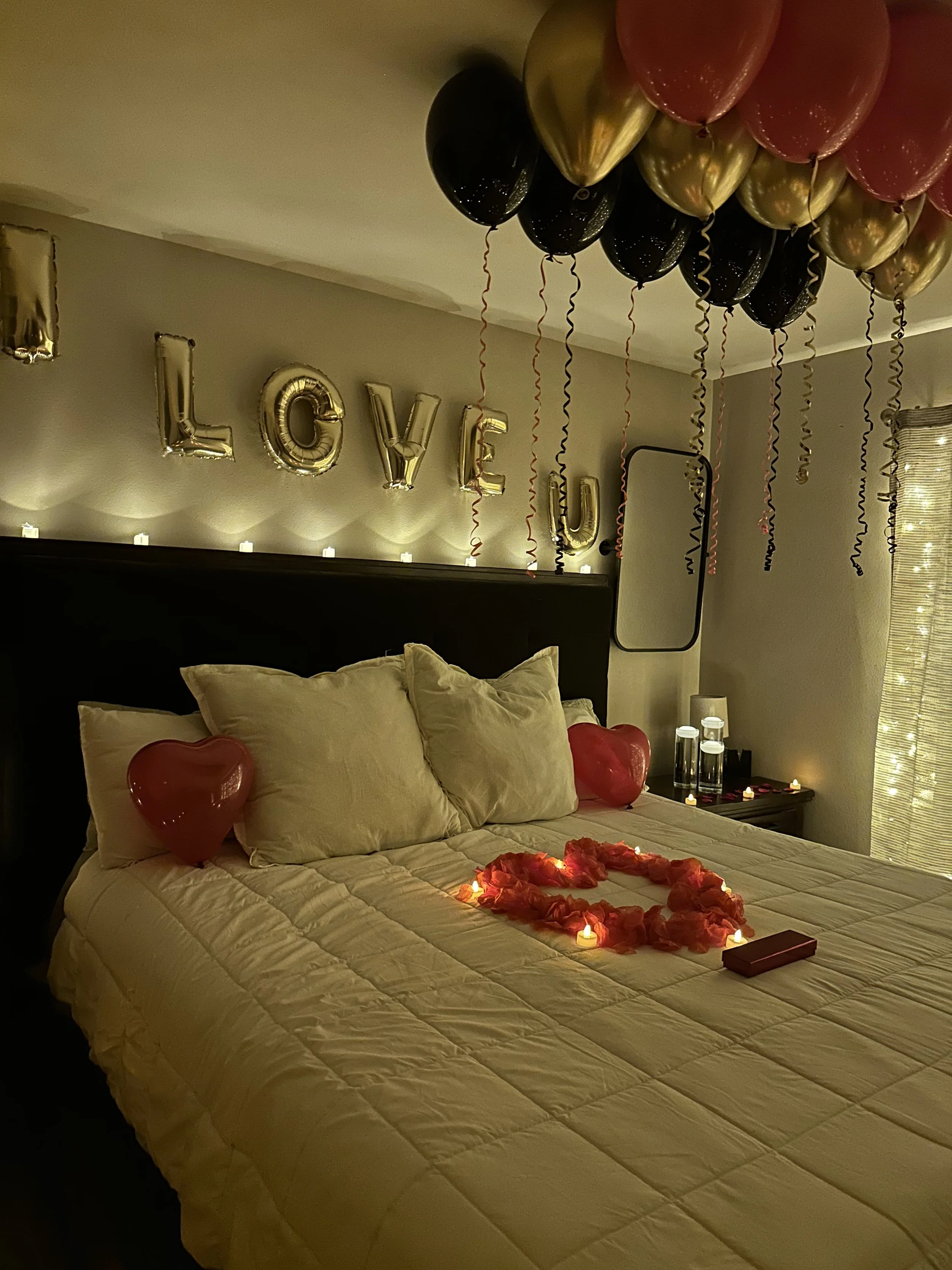 A bed decorated for a romantic occasion with heart-shaped balloons, candles, and rose petals. The wall behind has gold letter balloons spelling 'LOVE' and string lights, with balloons hanging from the ceiling.