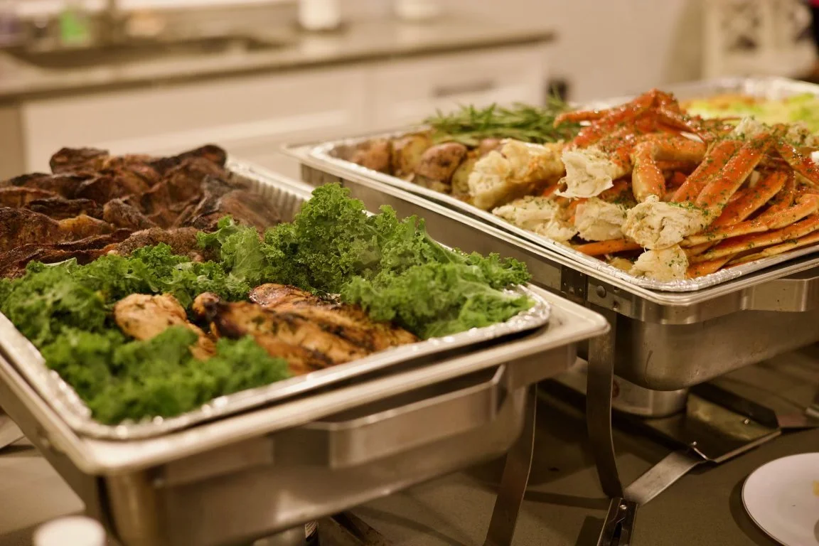 Buffet trays with grilled meats, fresh green lettuce, and seafood pasta topped with tomato sauce.