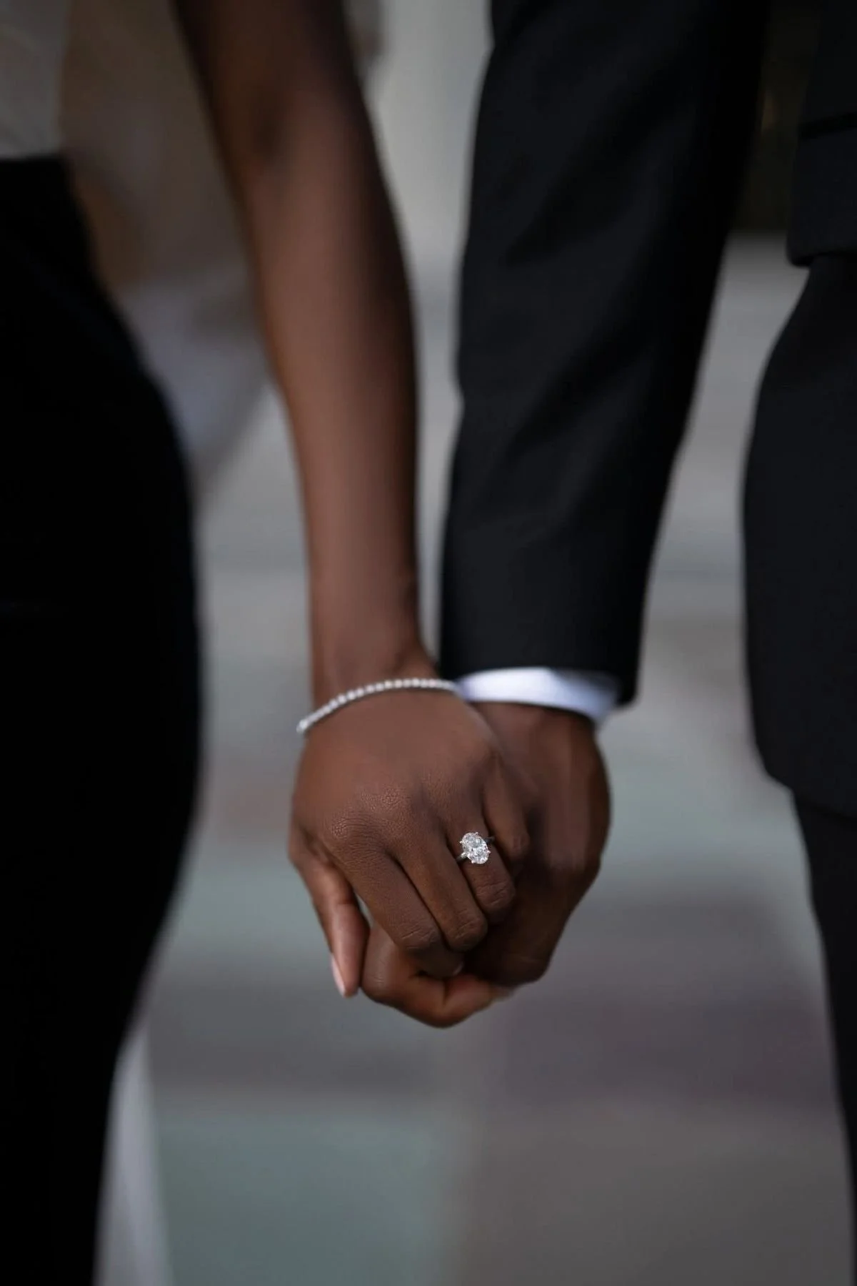 A couple holding hands, with the woman wearing a diamond engagement ring and a bracelet, and the man in a dark suit with white shirt cuffs visible.
