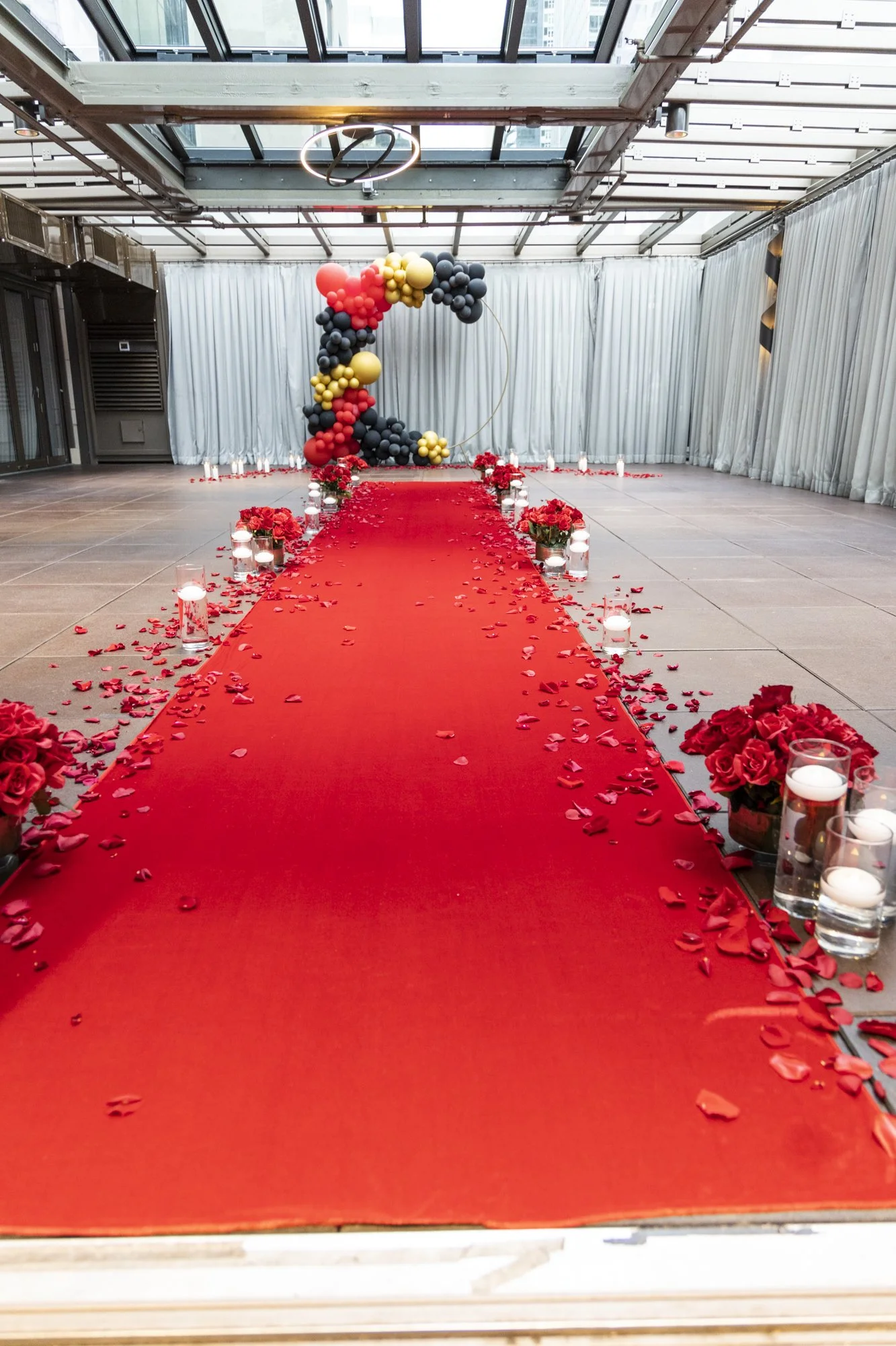 Indoor venue with a red carpet aisle decorated with rose petals, surrounded by candles and flower arrangements, leading to a balloon arrangement forming the number 2 in black, gold, red, and yellow balloons.