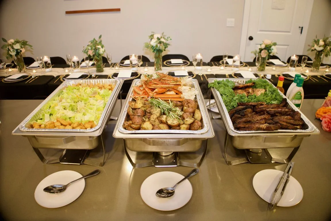 Buffet table with three chafing dishes containing salad, roasted vegetables and meats, garnished with leafy greens, set for a formal gathering.