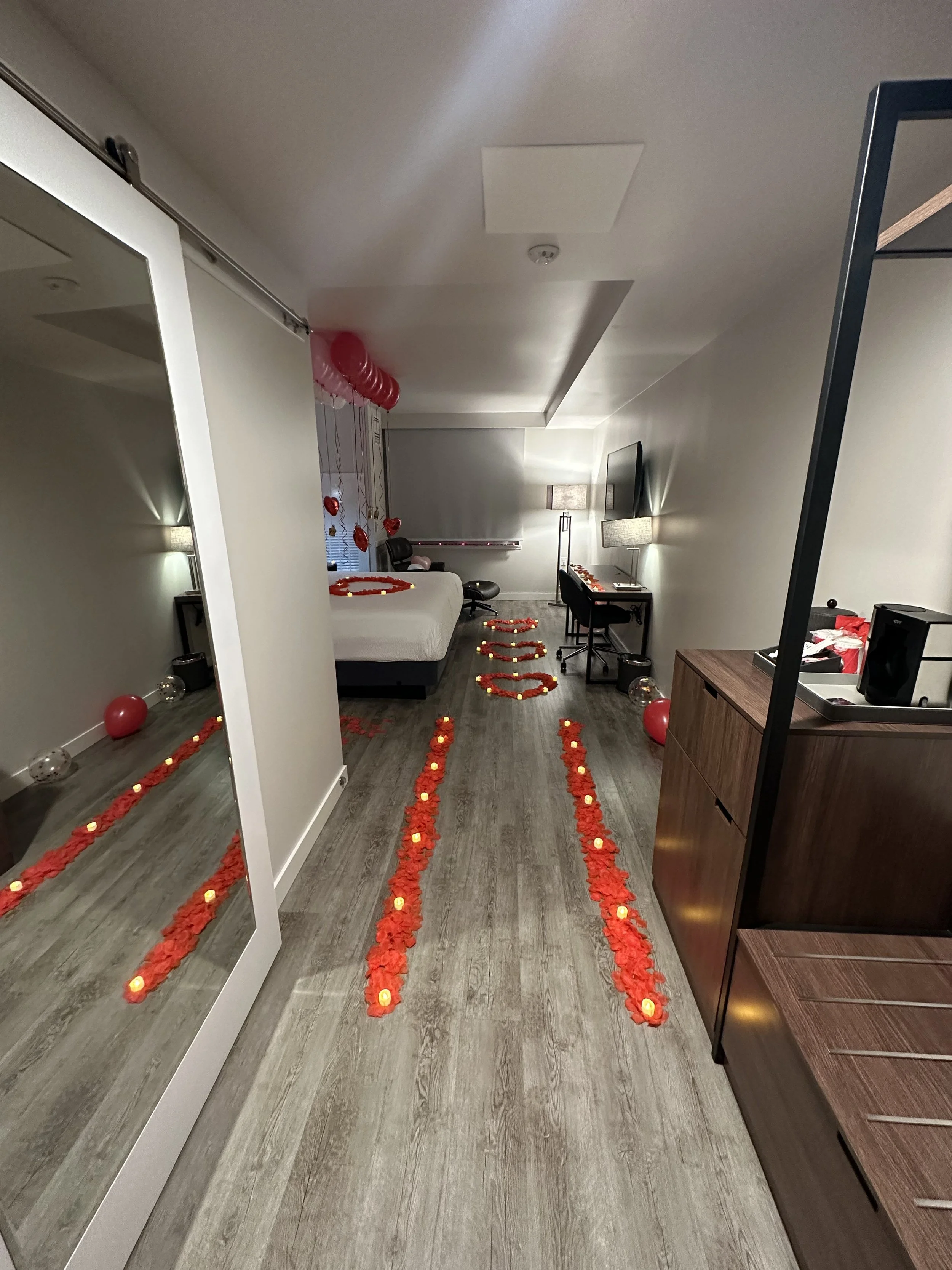 A hotel room decorated with red balloons, rose petals, and candles arranged in lines on the floor, creating a romantic pathway and heart shapes near the bed.
