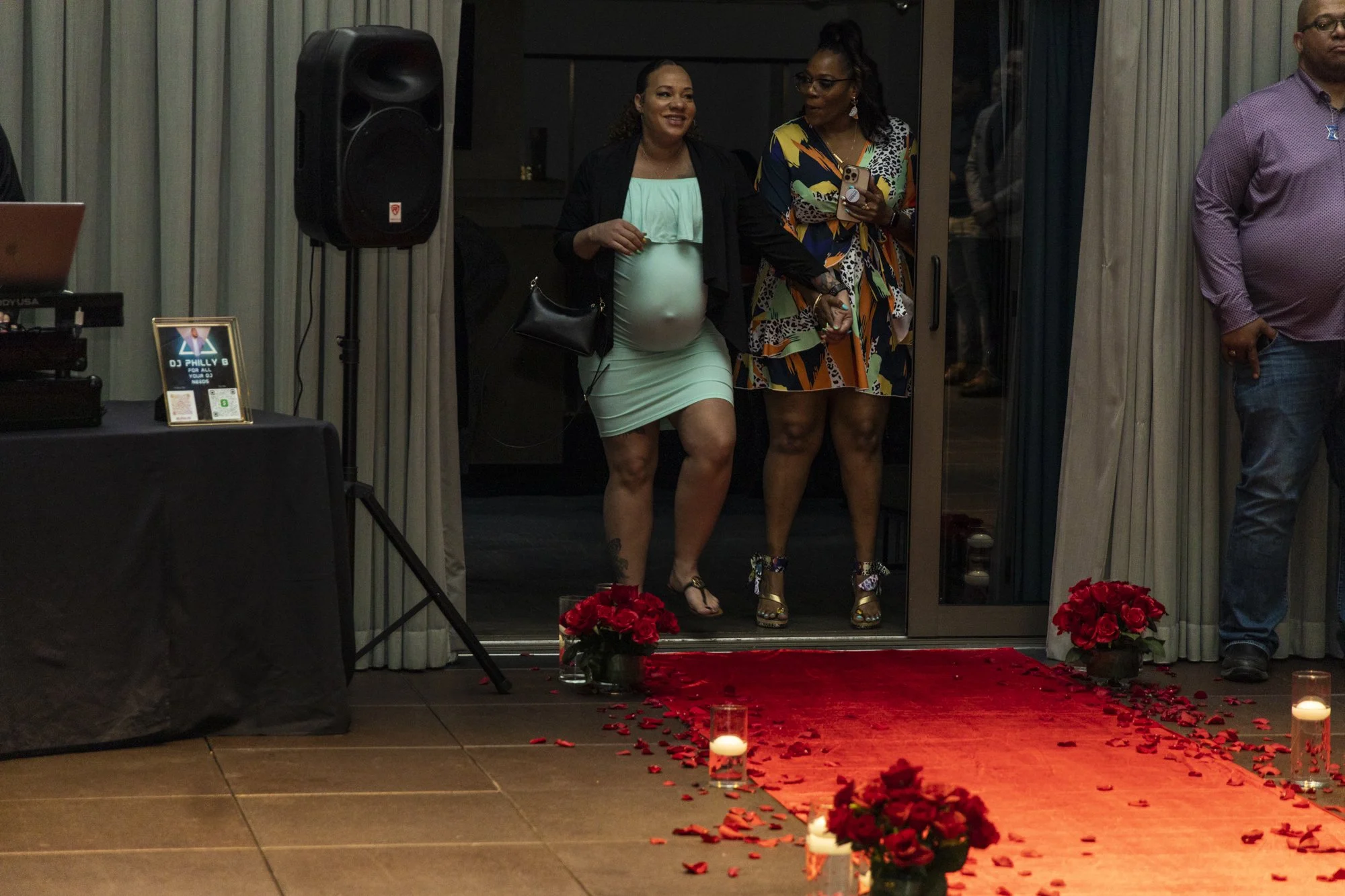 Two women standing at the entrance of a decorated indoor space with red roses, candles, and rose petals, possibly at a celebration or event.