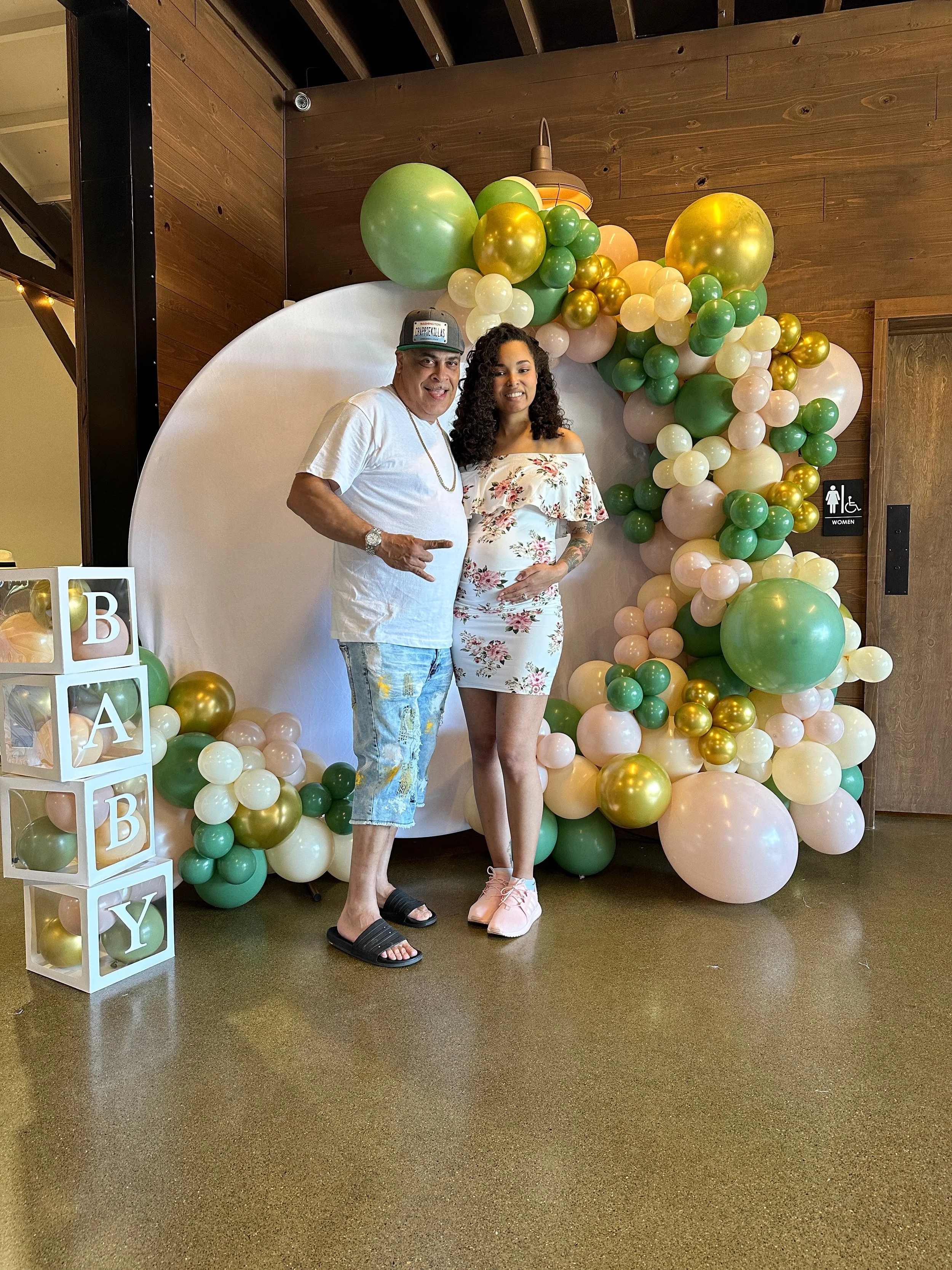 A man and woman posing in front of a balloon backdrop with shades of green, white, cream, and gold balloons. The man wears a white t-shirt, ripped shorts, a cap, and sandals, and the woman is dressed in a white floral dress and pink shoes.