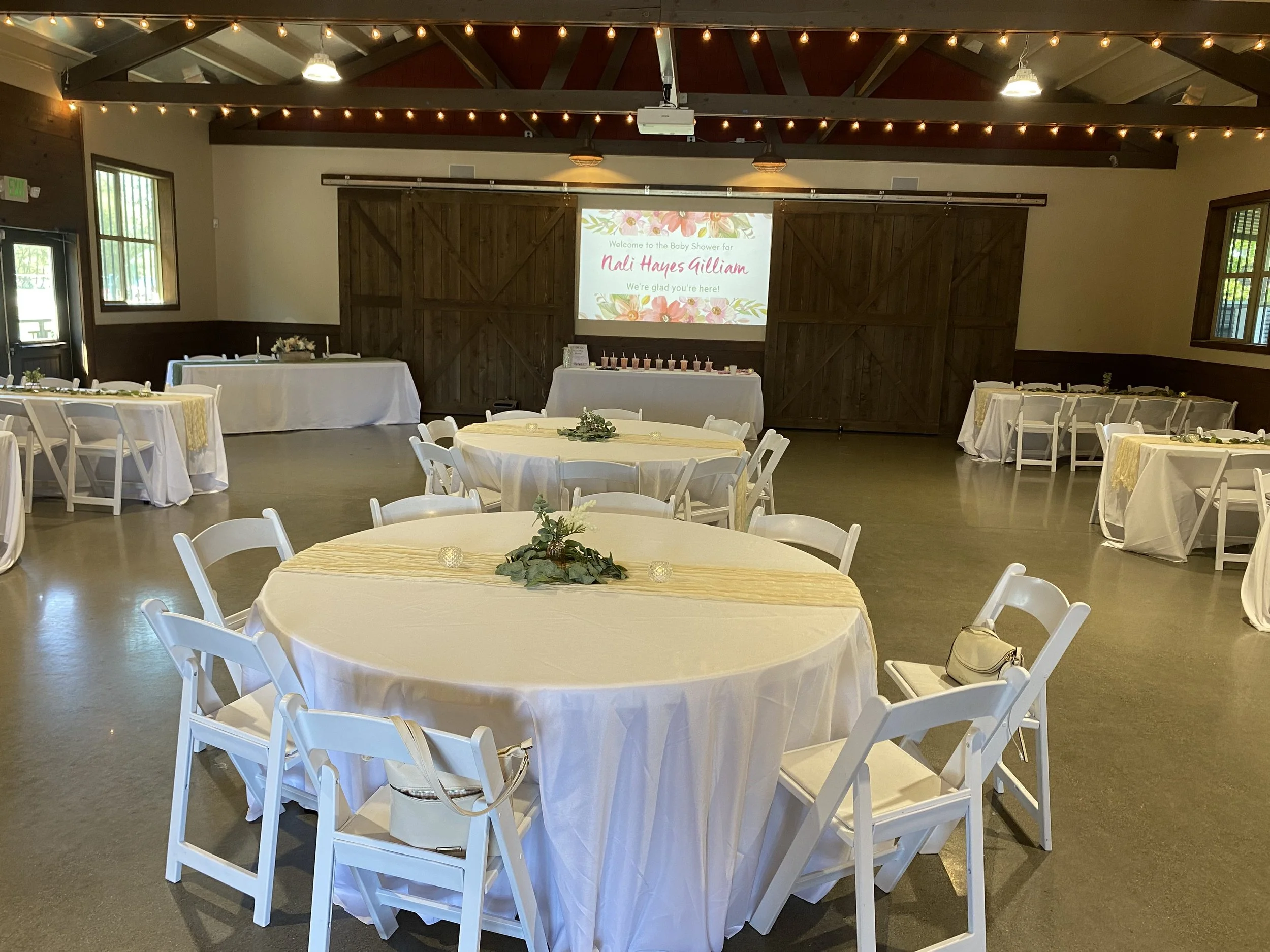 Event hall decorated for a baby shower with round tables and white chairs, a projector screen with a welcome message for Nali Hayes Gillam, and a table with pink candles.