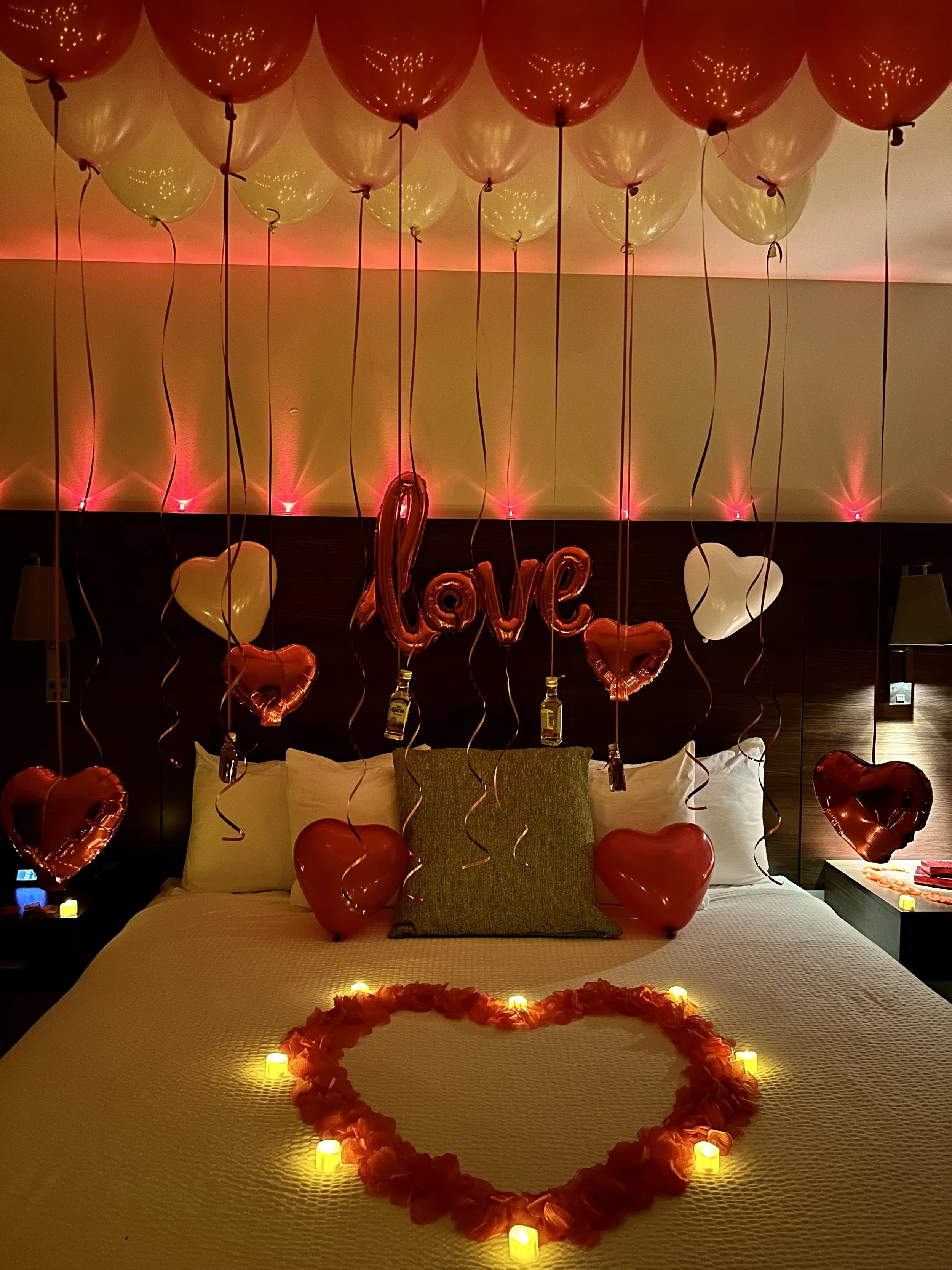 Decorated hotel bed for a romantic occasion with heart-shaped balloons, candles, and a bedspread, featuring balloons spelling 'love' and a heart-shaped floral arrangement.