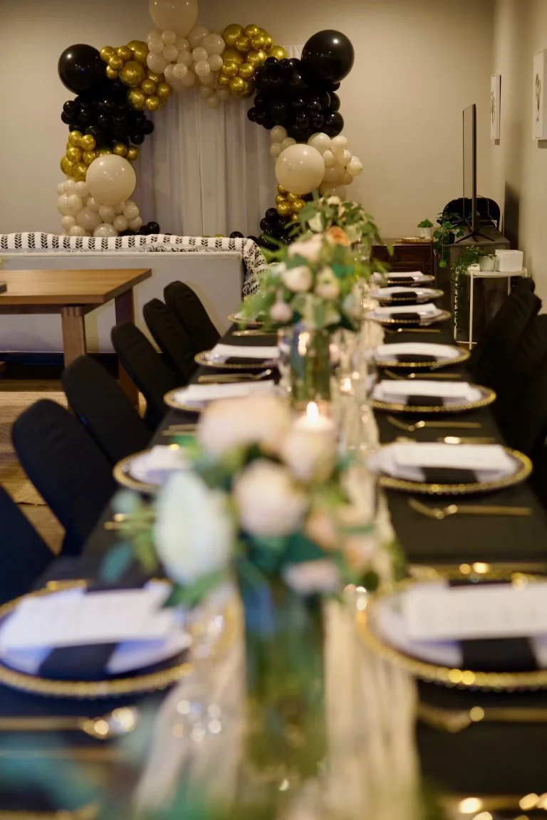 A decorated dining table with floral centerpieces, black and gold plates, and black chairs, with a balloon arch in black, white, and gold in the background.