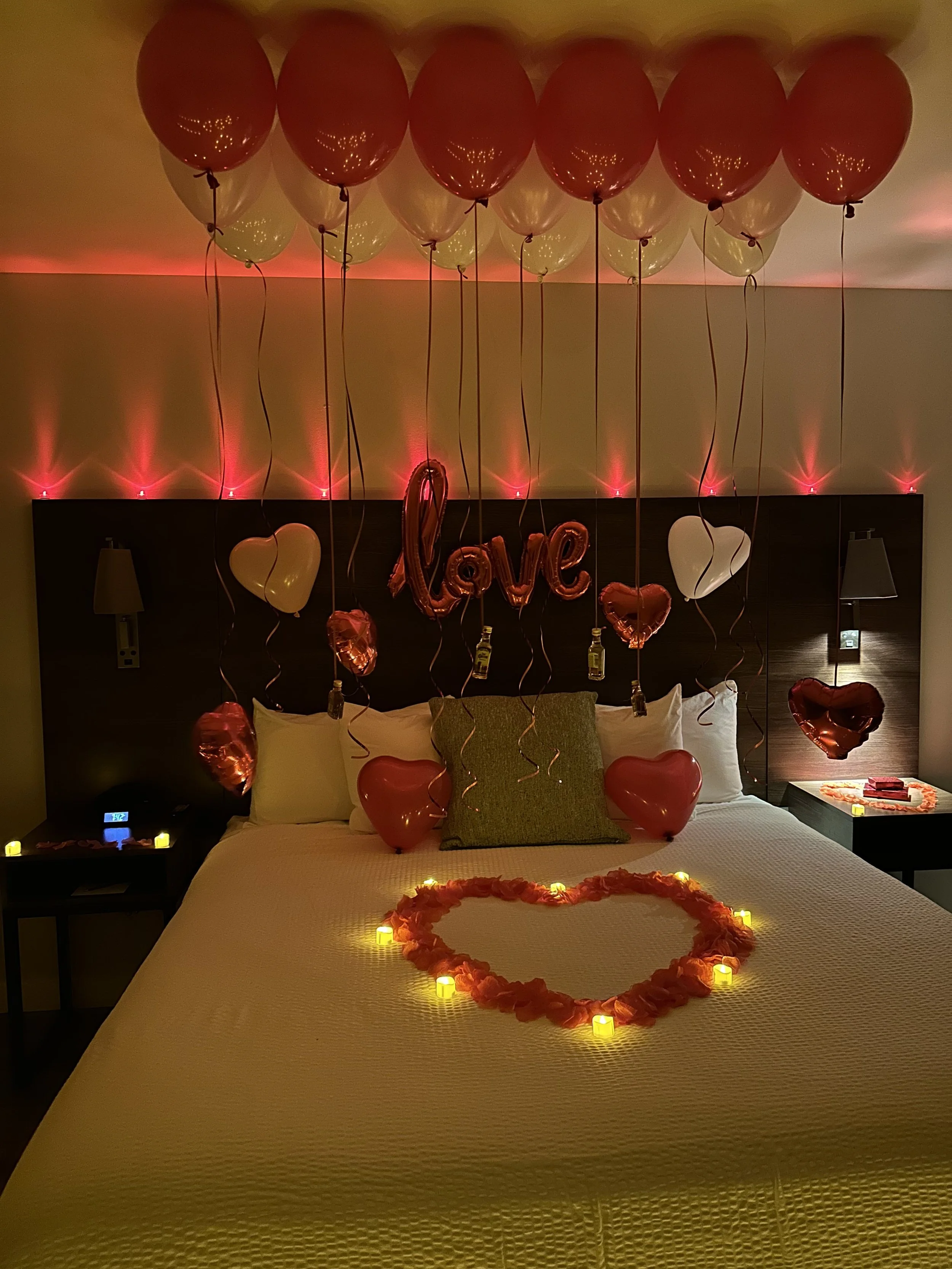 A hotel room decorated for a romantic occasion with red and white balloons hanging from the ceiling, illuminated candles forming a heart shape on the bed, and balloons and lights spelling out the word "love" on the headboard.